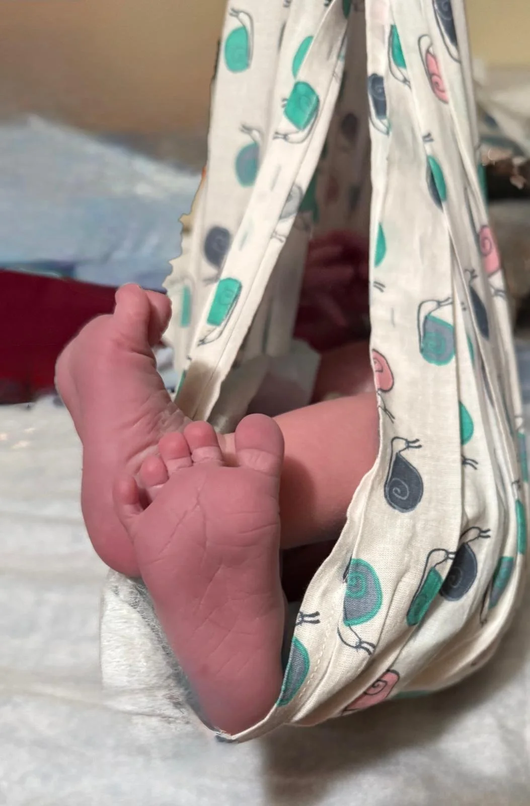A newborn baby is sleeping inside a fabric tent with a colorful snail and leaf pattern. The baby is lying on its side, with tiny fingers clenched near its face. The fabric tent is partly open, revealing the baby's face and hand.