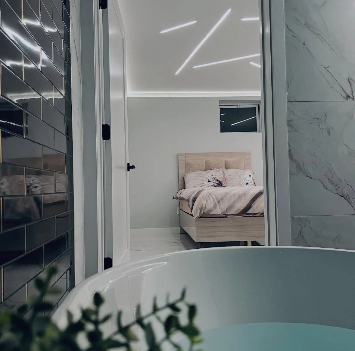 A bathroom with black subway tiles, a partial view of a bathtub, and a view into a bedroom with a bed and a small window.
