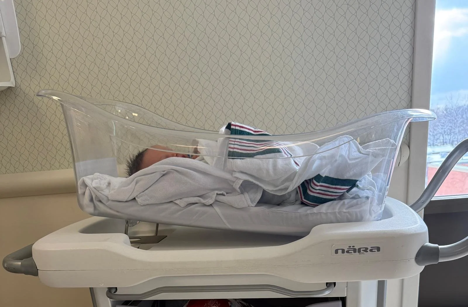 Newborn baby in an incubator in a hospital room, resting inside the clear plastic enclosure.