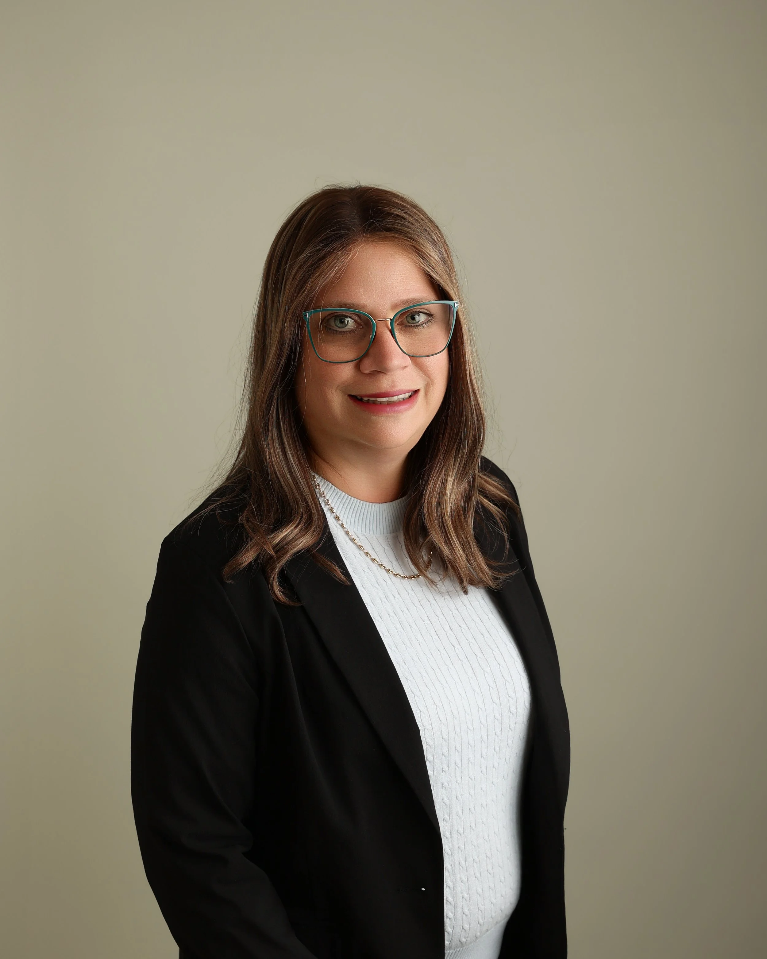 A woman with brown hair wearing glasses, a white sweater, and a black blazer, smiling in front of a plain light background.
