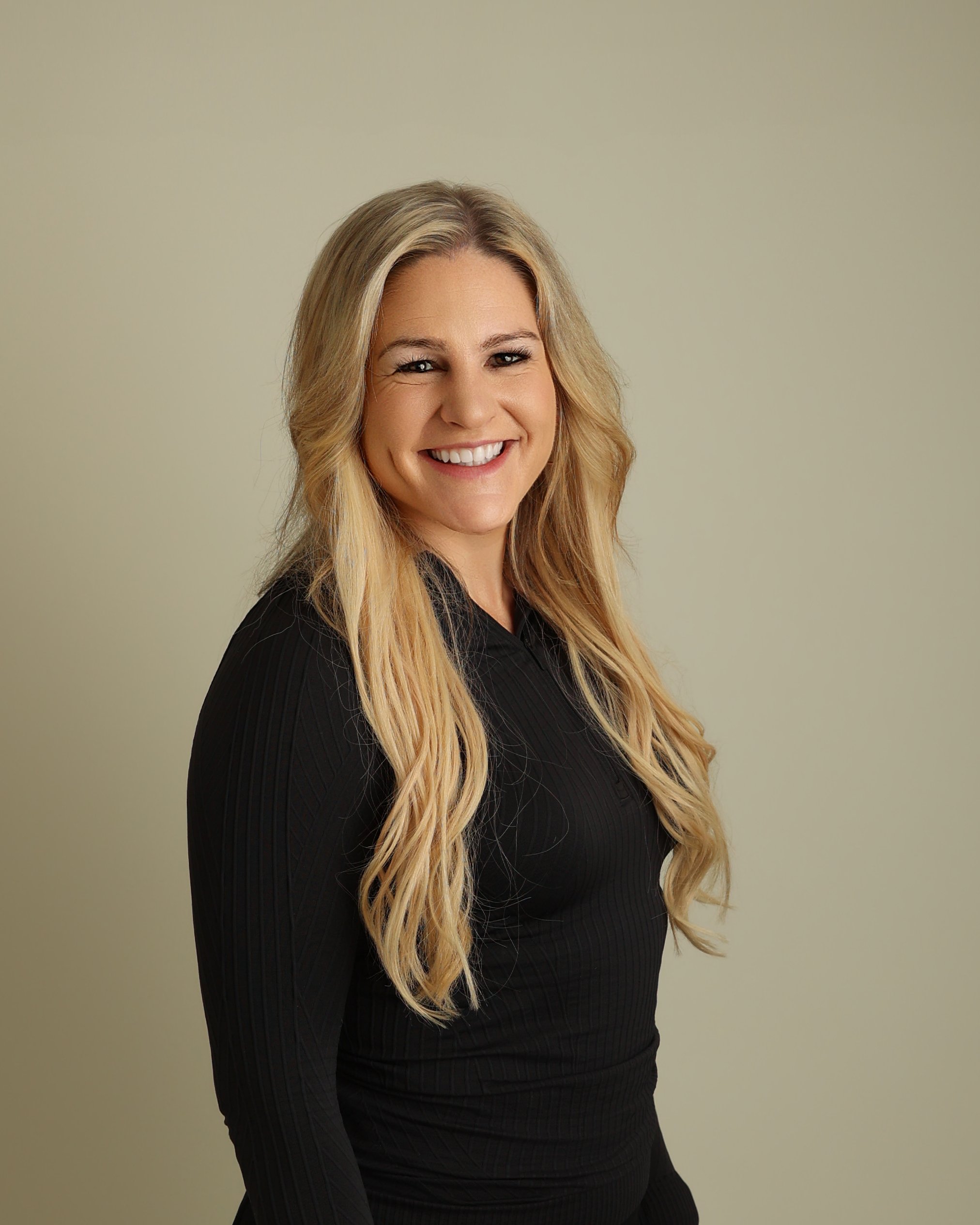 A woman with long blonde hair smiling, wearing a black long-sleeve top, standing against a plain light-colored background.