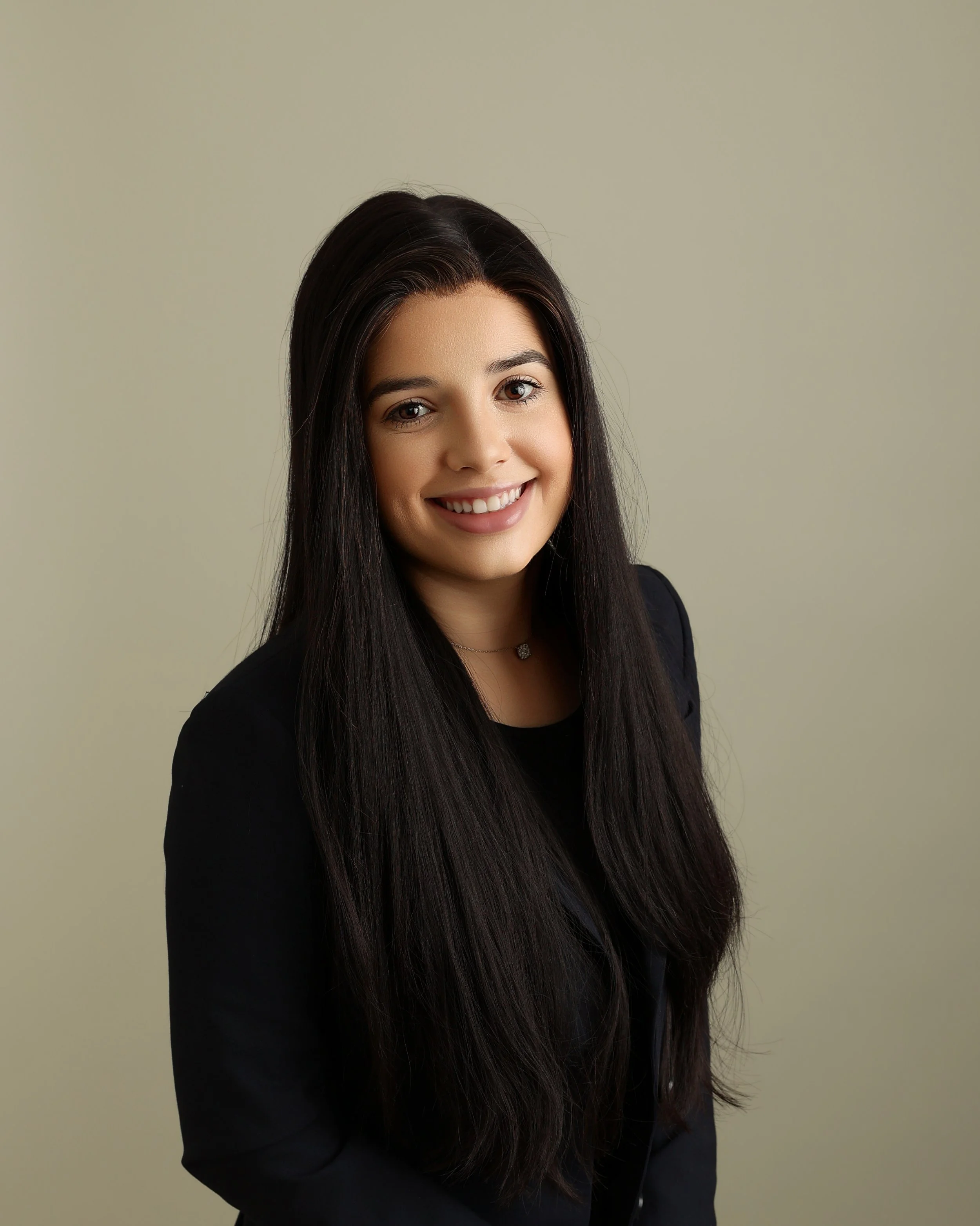 A woman with long dark hair smiling, wearing a black top and a necklace, standing against a plain beige background.