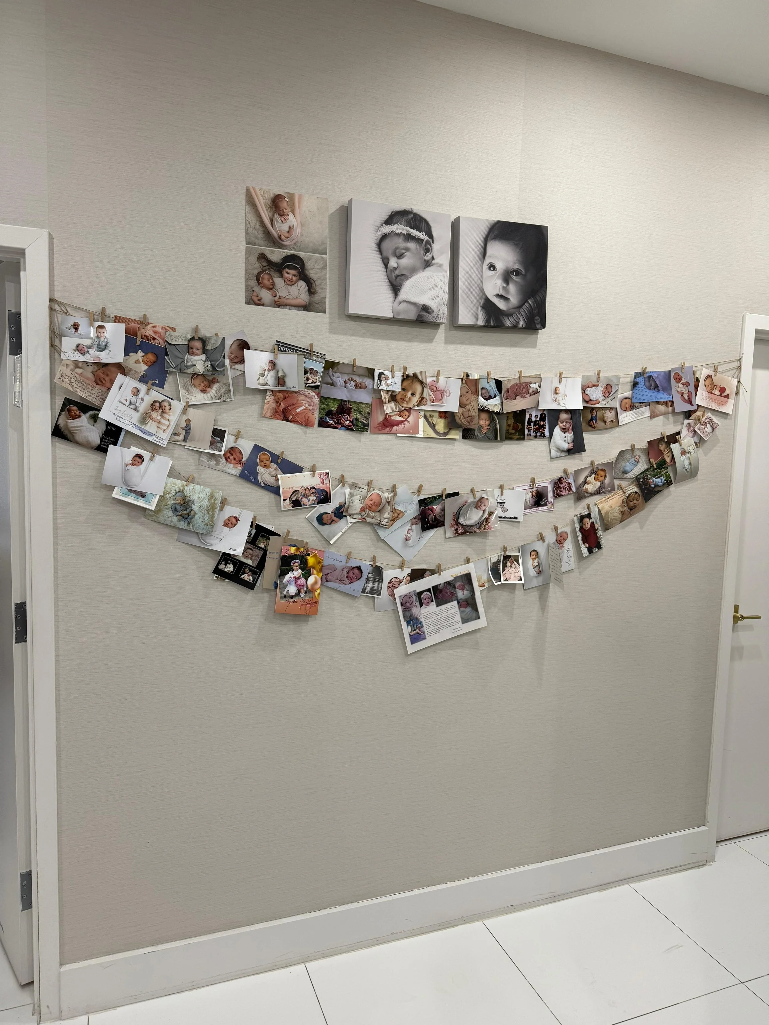 Display of many printed photographs of babies and children hanging on a wall with clothespins, including black and white photos of a sleeping baby and a child looking at the camera, and additional color photos.