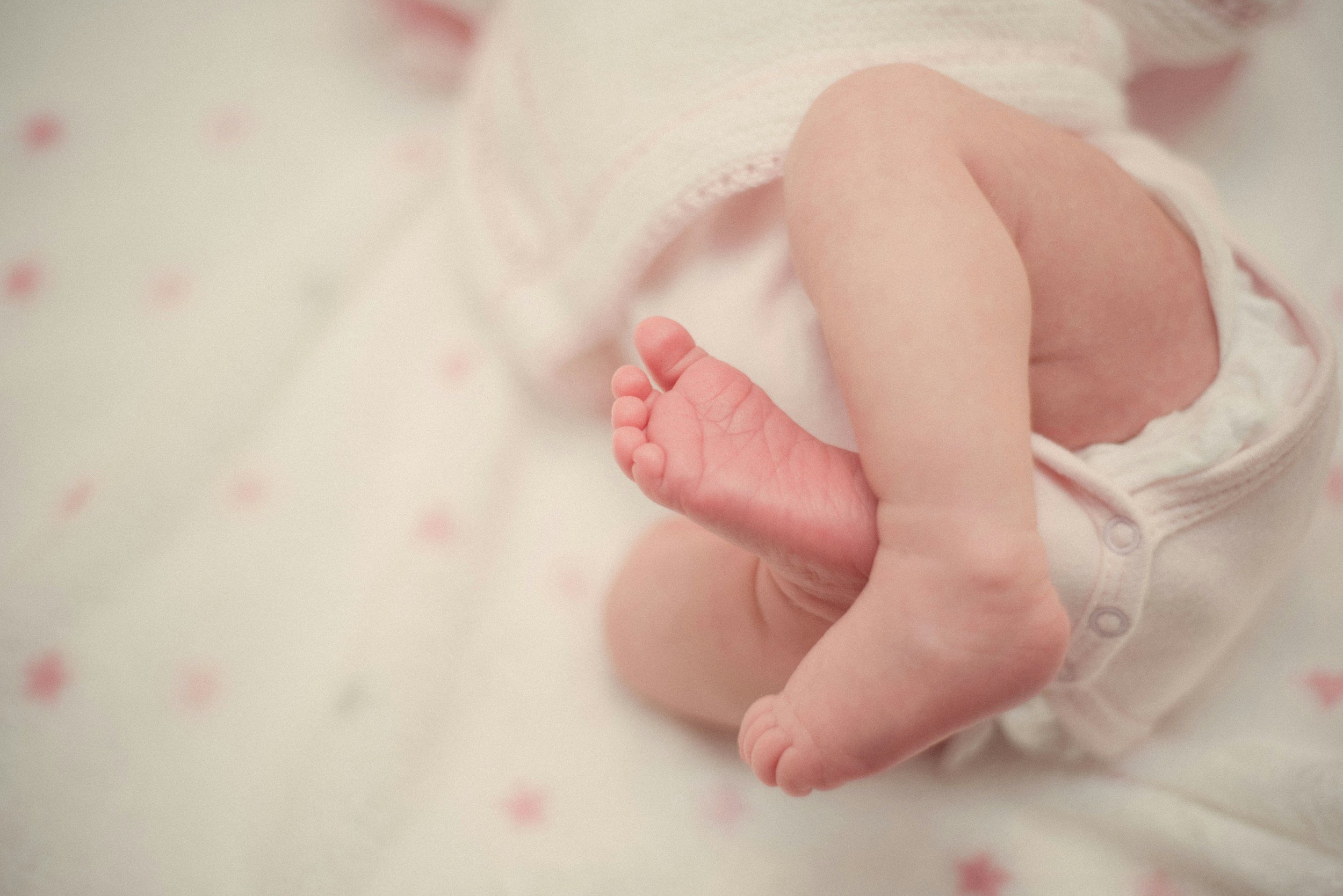 Close-up of a newborn baby holding an adult's finger, with the baby wearing a white outfit with small pink details.