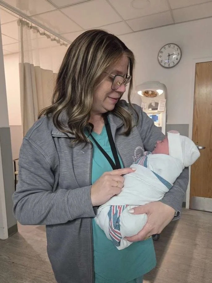 Woman in scrubs and glasses holding a newborn baby in a hospital room.