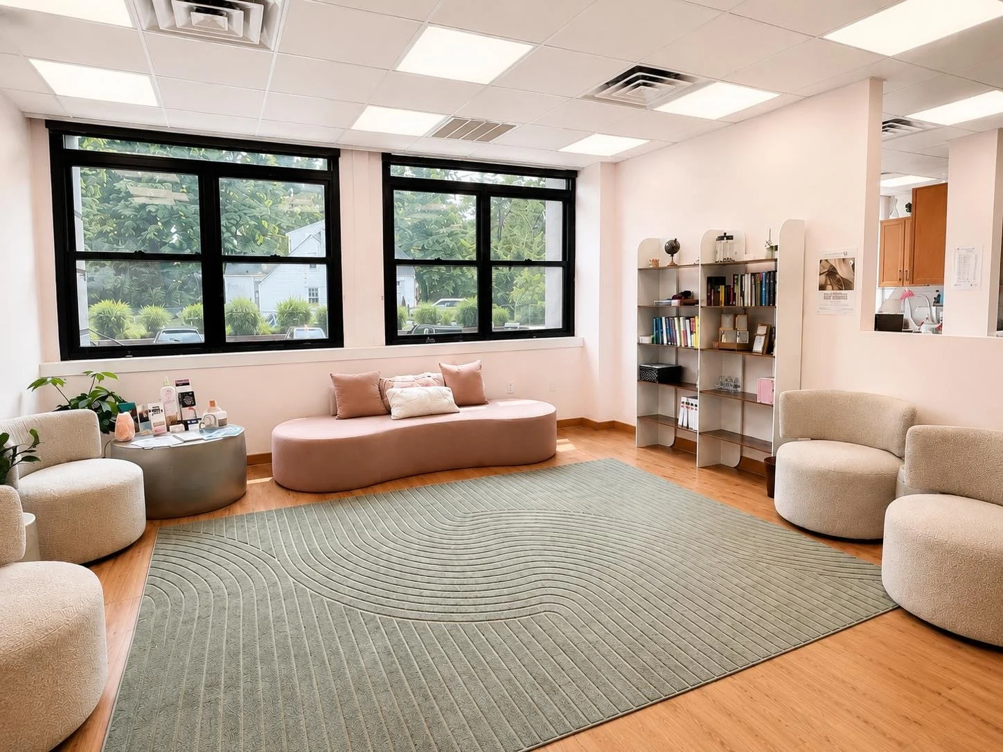A spacious waiting room with large windows, beige and pink seating, a green patterned rug, a gray side table with skincare products, and a white bookshelf filled with books and decorative items.