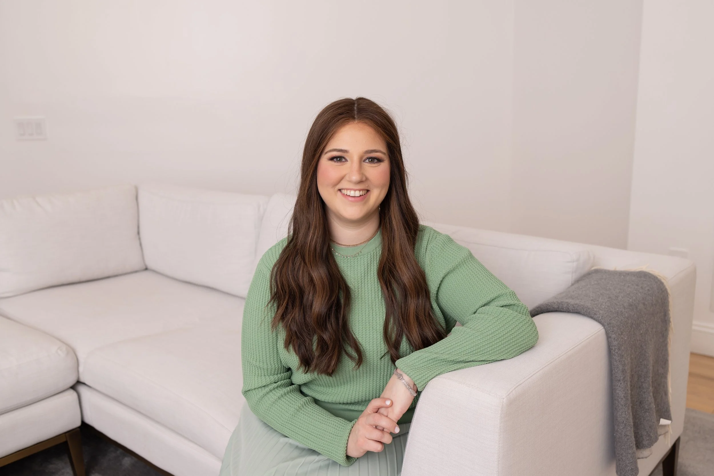A smiling woman with long brown hair and light makeup sitting on a white sofa in a bright room with white walls, wearing a green sweater, with her arm resting on a white chair.