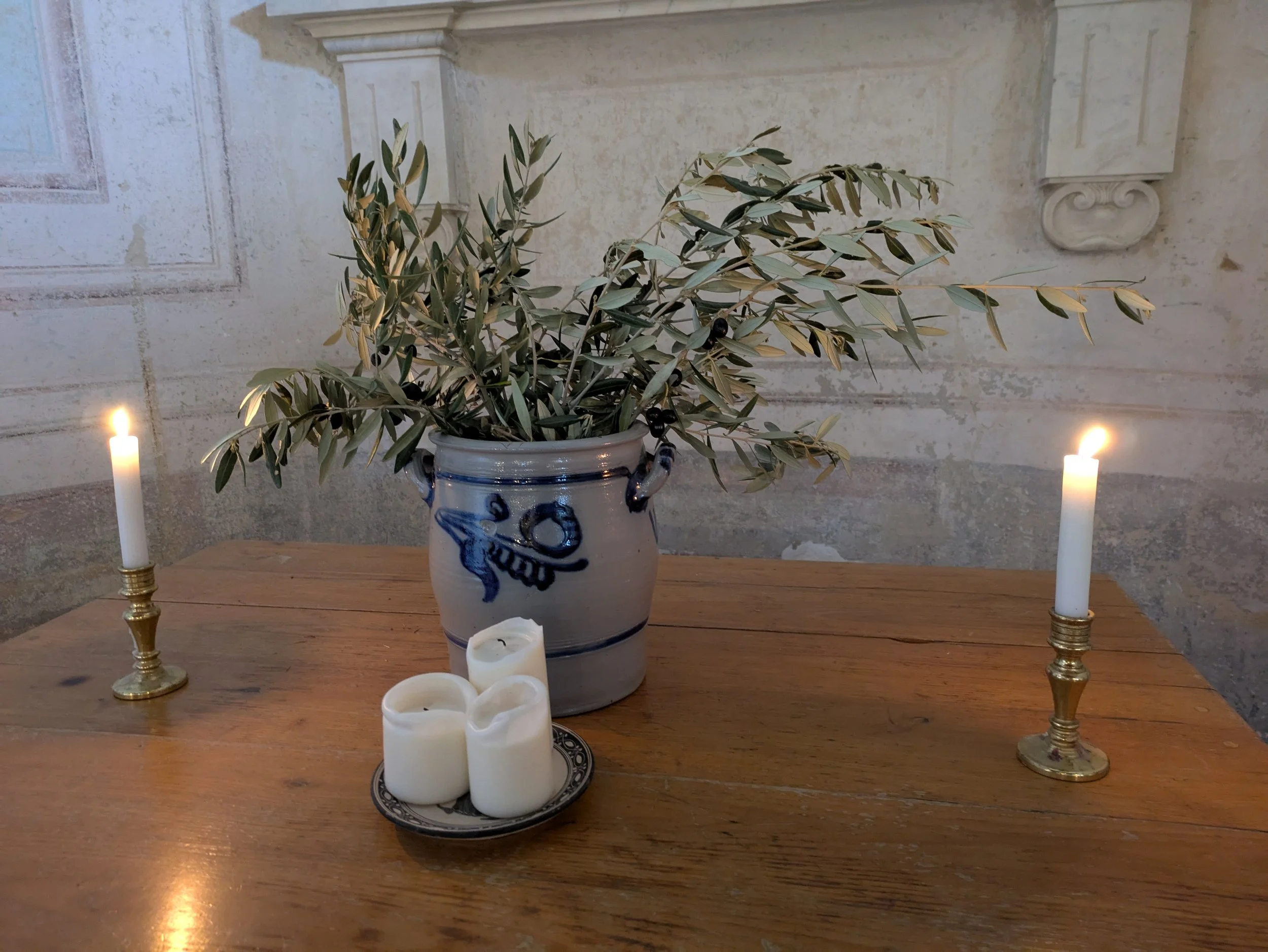 A table with a blue and white ceramic vase holding green and yellowish olive branches, surrounded by several white candles and two brass candle holders, with a light-colored textured wall in the background.