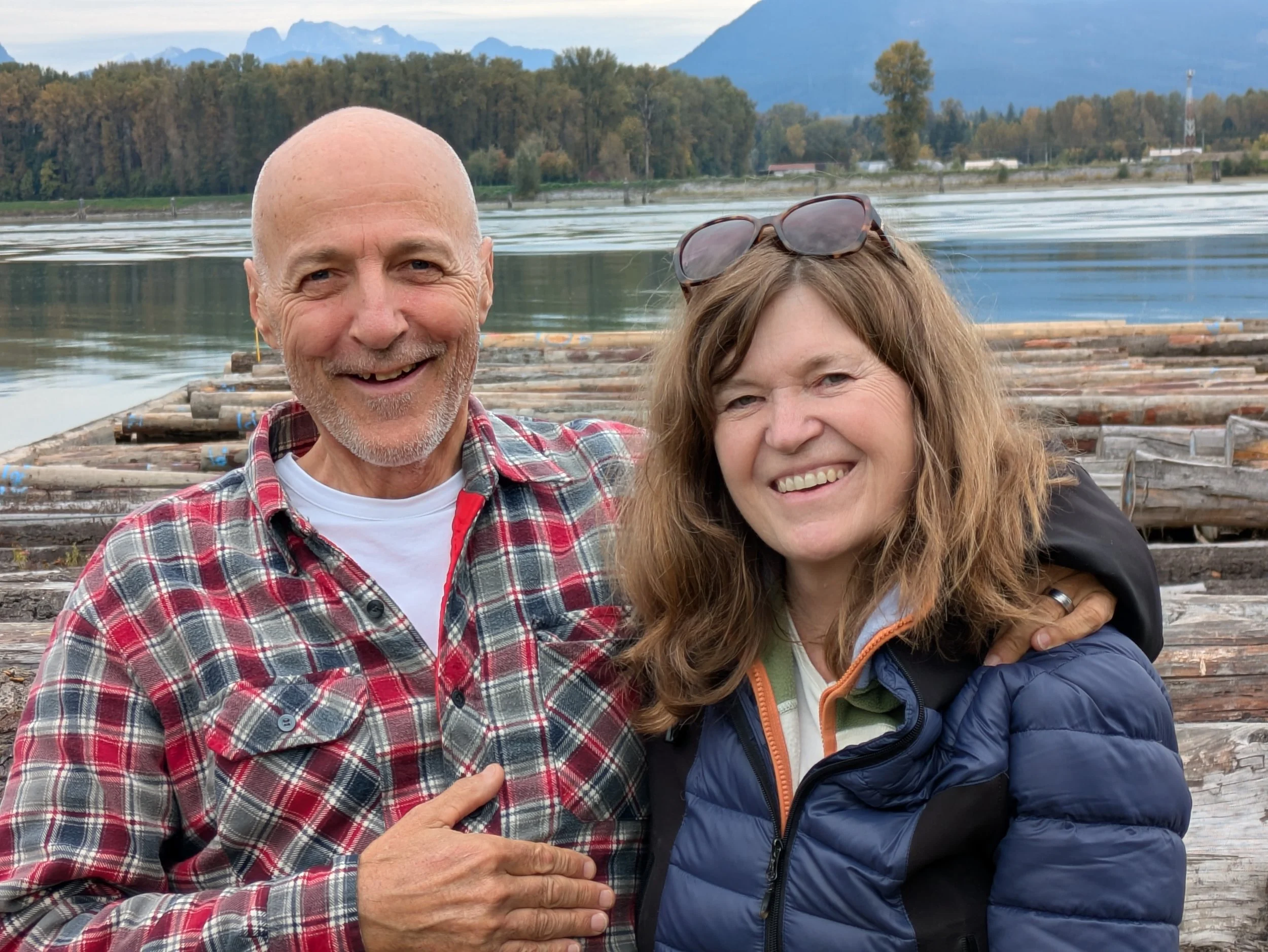 A smiling older man with a bald head and gray beard wearing a red plaid shirt and a woman with long curly hair wearing sunglasses on her head and a blue jacket, both standing outdoors near a body of water with mountains and trees in the background.