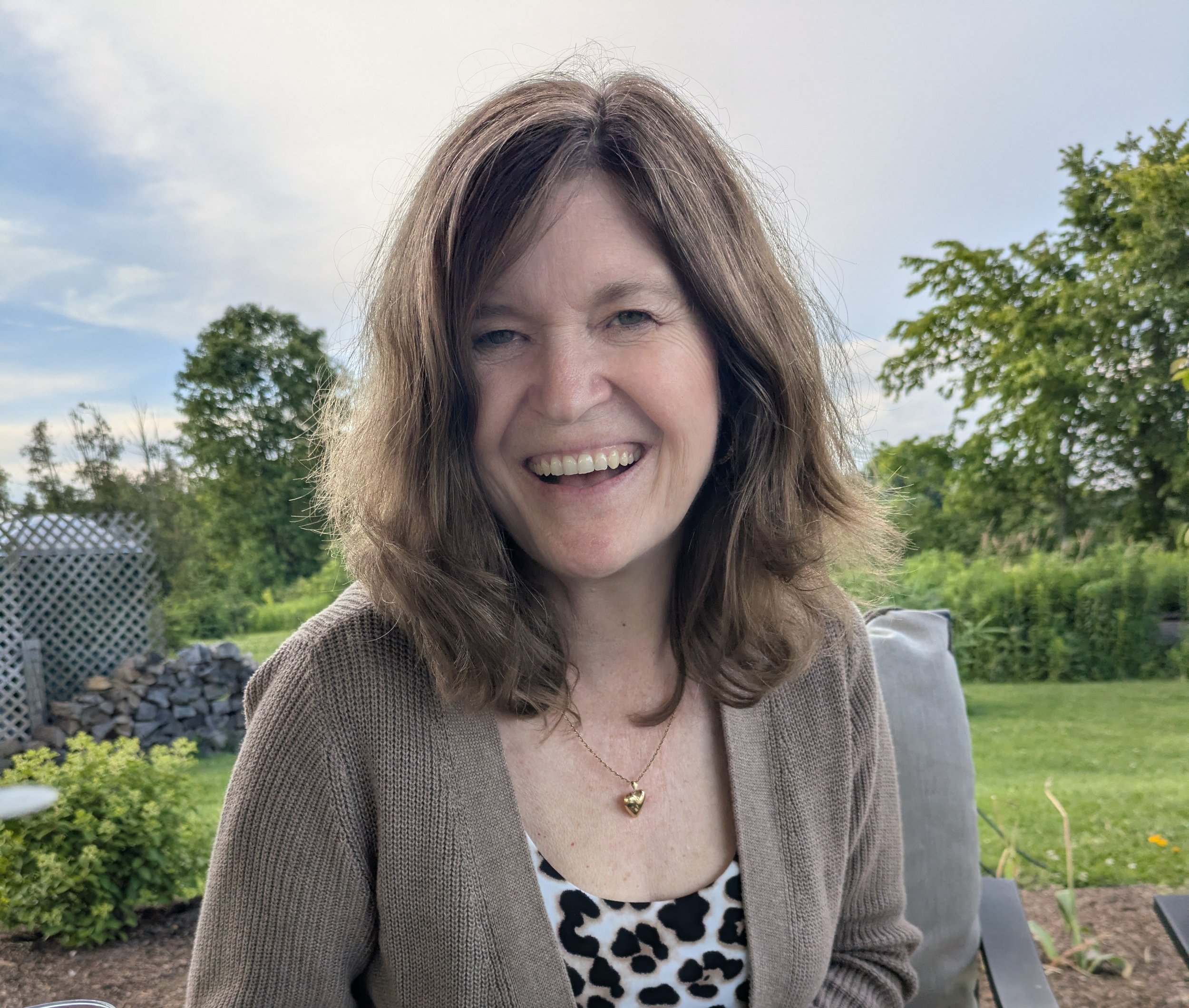 A woman with long brown hair smiling outdoors in a backyard with trees and a cloudy sky in the background.