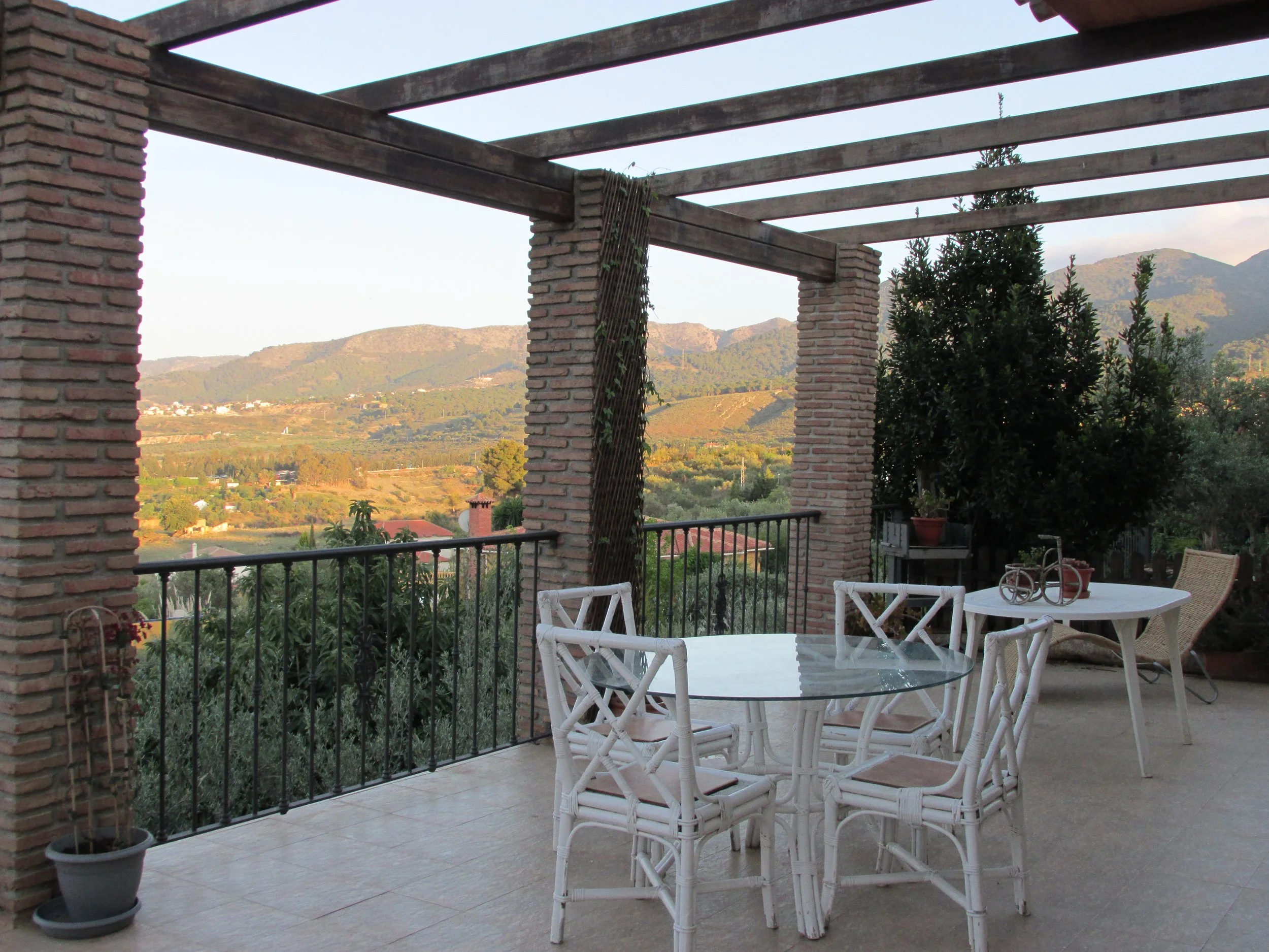 An outdoor terrace with a round glass table and white chairs, surrounded by brick columns and a black metal railing. There are potted plants on either side of the terrace, and in the background, there are green hills and mountains under a clear sky.