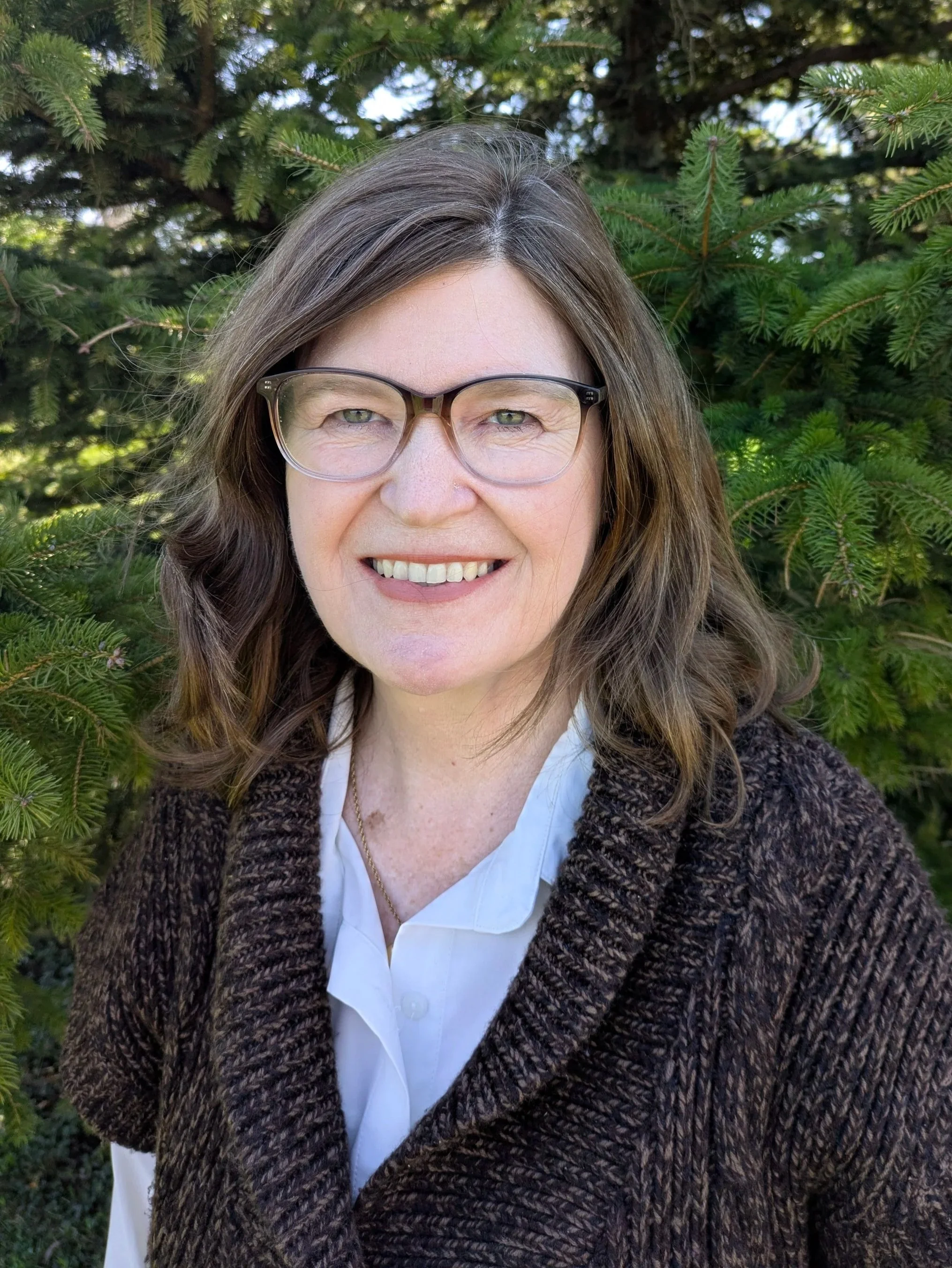 A woman with glasses smiling outdoors, in front of green pine branches.