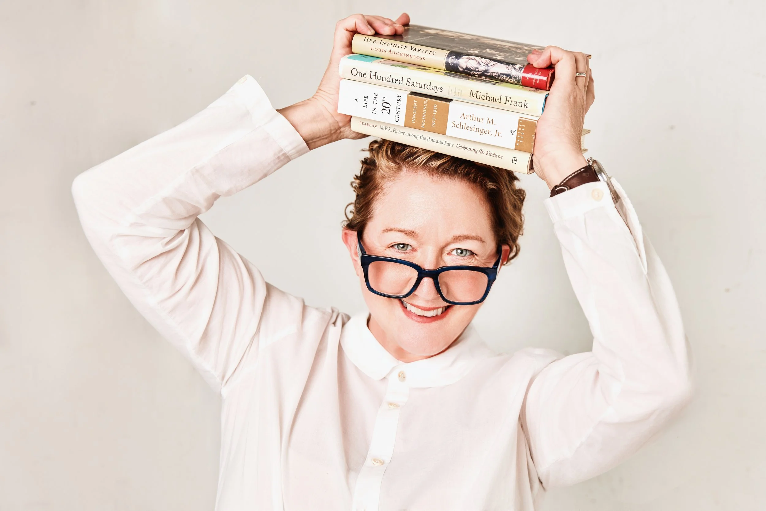 A person with curly hair and glasses holding a stack of books on their head, smiling at the camera.