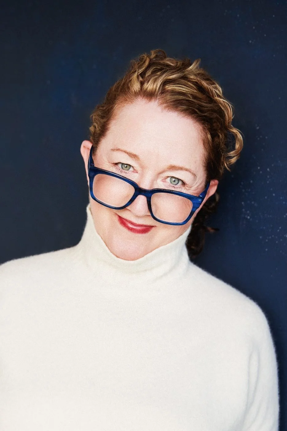 A woman with short, curly reddish-brown hair, wearing a white turtleneck sweater and large blue glasses, smiling against a dark, starry background.