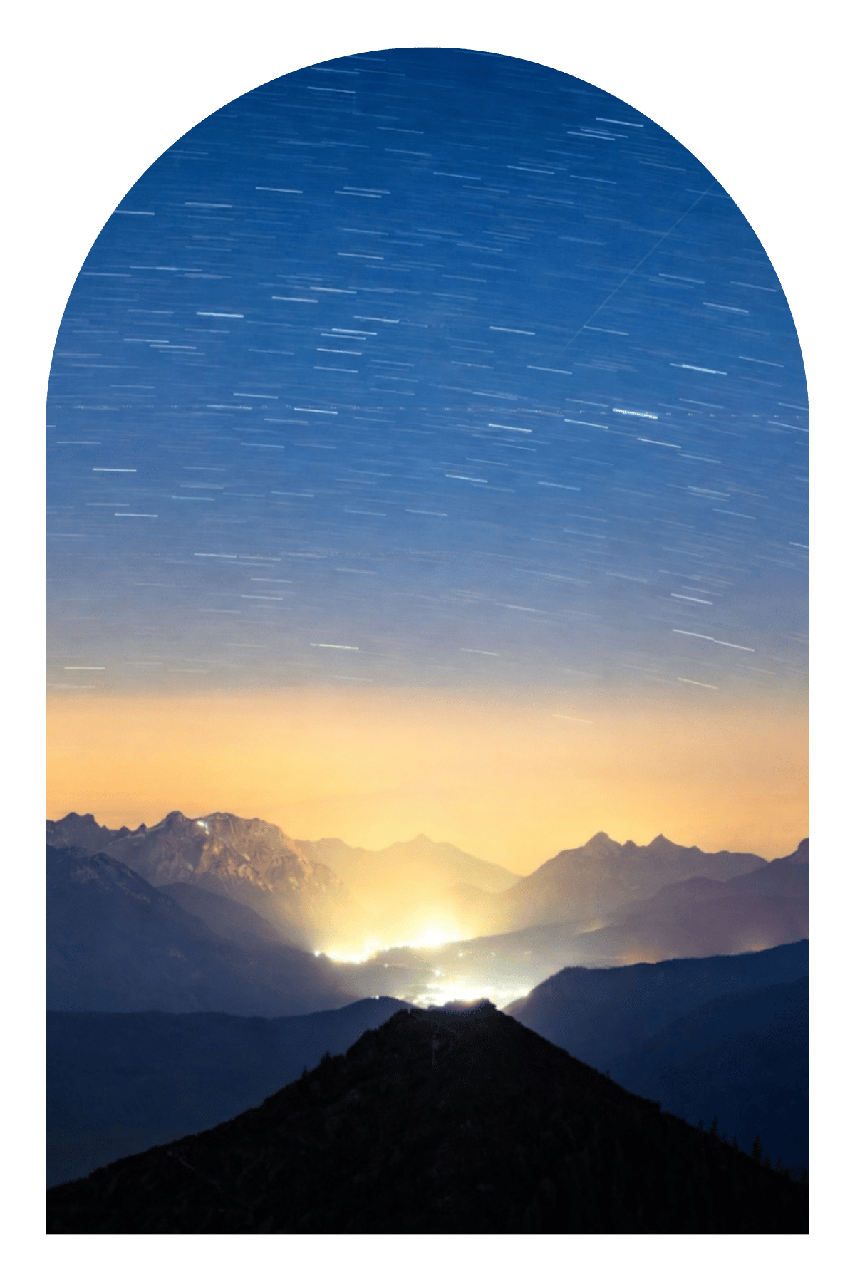 Star trails sweeping across a deep blue night sky above a peaceful mountain valley, with warm golden light glowing along the distant horizon.