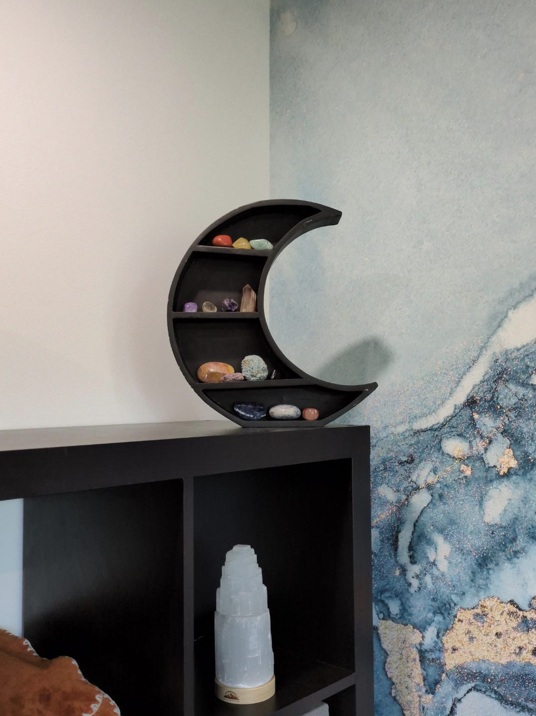 Black crescent moon shelf displaying colorful crystals and stones against a textured blue and gold wall.