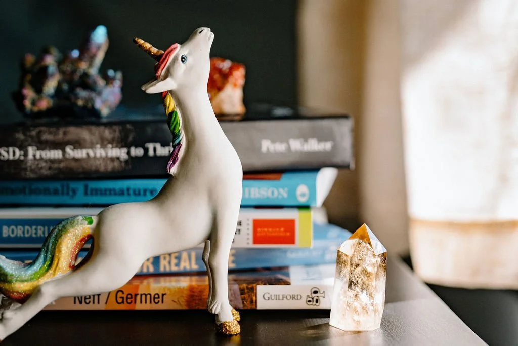 A playful white unicorn figurine with a rainbow mane, glittery tail, and gold hooves displayed in front of a collection of books in Omy Golden's therapy office.