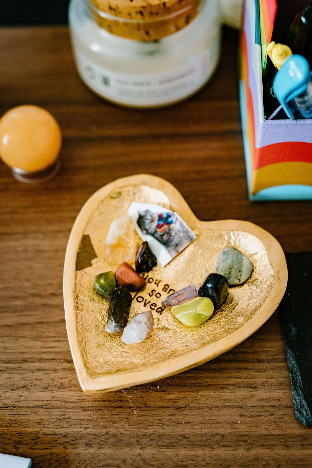 A gold heart-shaped dish filled with colorful healing crystals and small keepsakes, featuring the words “You are so loved,” resting on a wooden table.