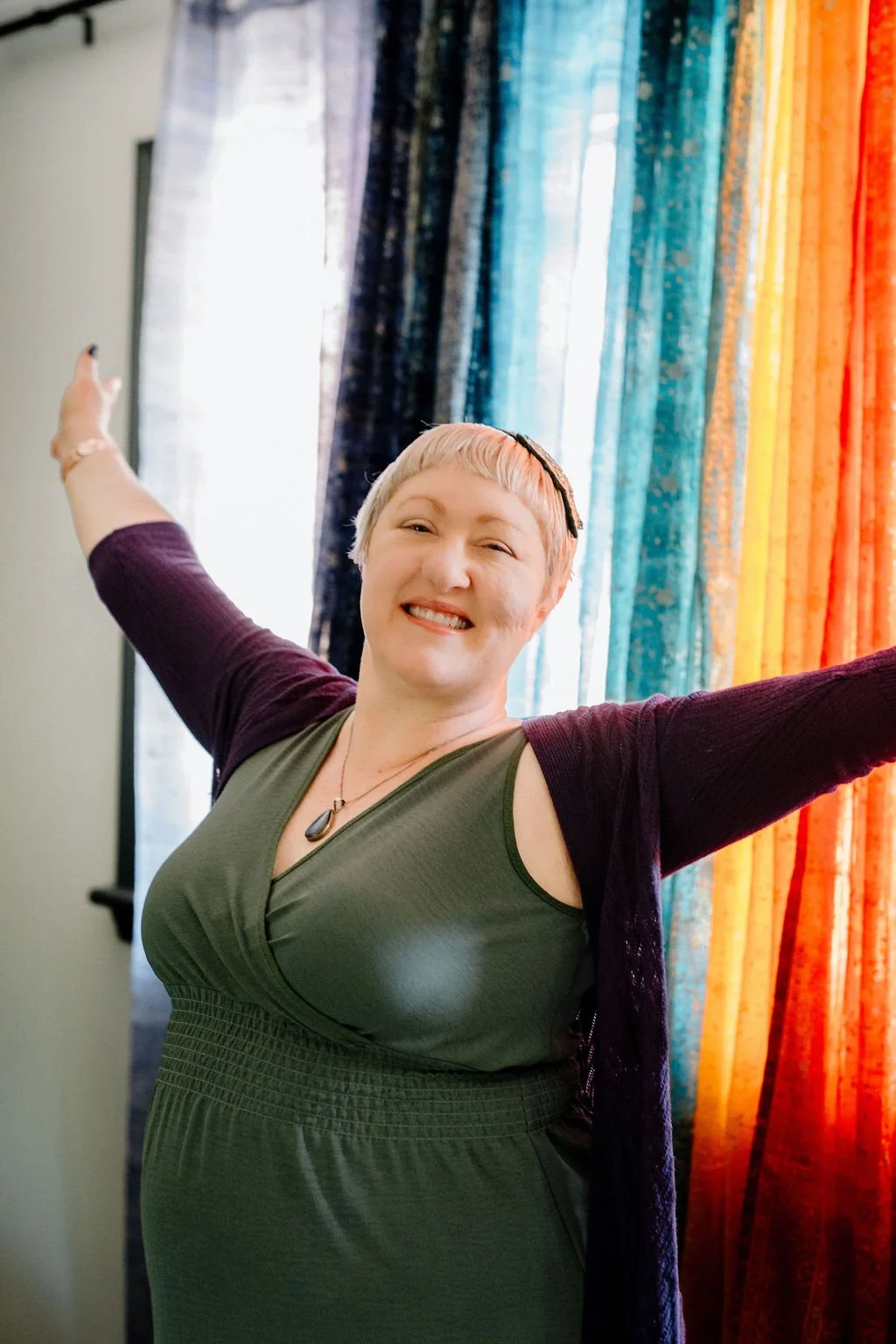 Omy Golden smiling with her arms outstretched in front of vibrant rainbow-colored curtains.