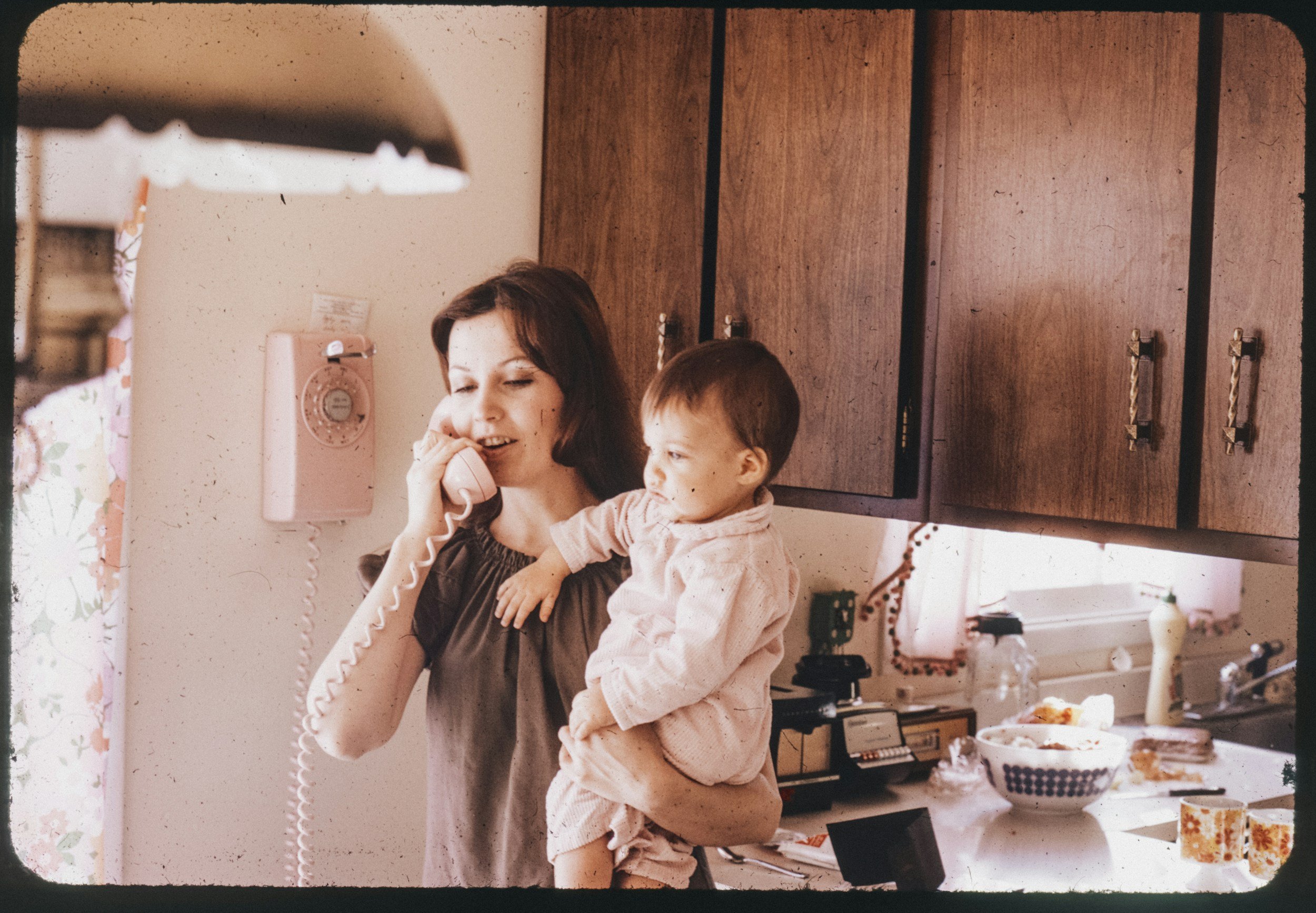 A woman talking on a pink rotary phone while holding a child in a kitchen. The woman has dark hair and is wearing a dark top, the child has short brown hair and is wearing a light-colored shirt. The kitchen has wooden cabinets, a pink rotary phone on the wall, and various kitchen items on a counter.