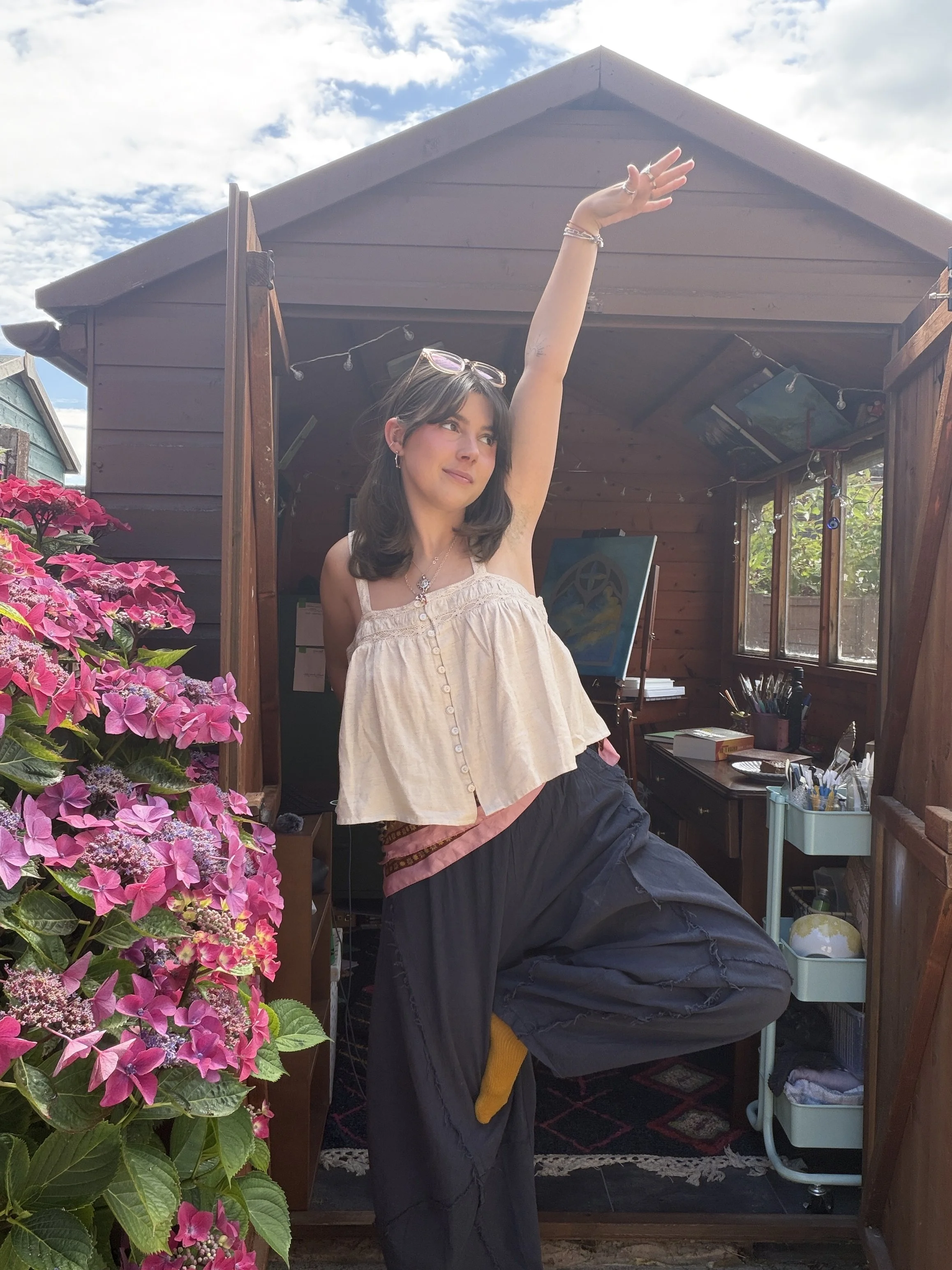 A woman standing in a small wooden room or garden shed, raising her right arm into the air. She has dark hair, wears glasses on her head, earrings, and a necklace, and is dressed in a sleeveless cream-colored top and loose dark pants. There are pink hydrangea flowers in the foreground on the left, and a shelf with art supplies and paintings inside the shed.