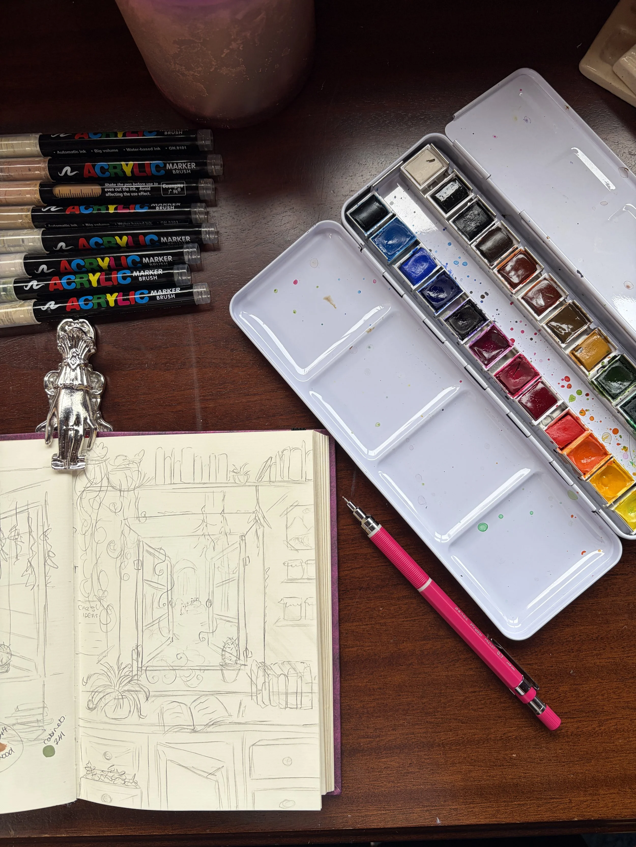 Open sketchbook with a detailed indoor scene drawing, watercolor paints in a case, a pink pen, and acrylic paint markers on a dark wooden table.