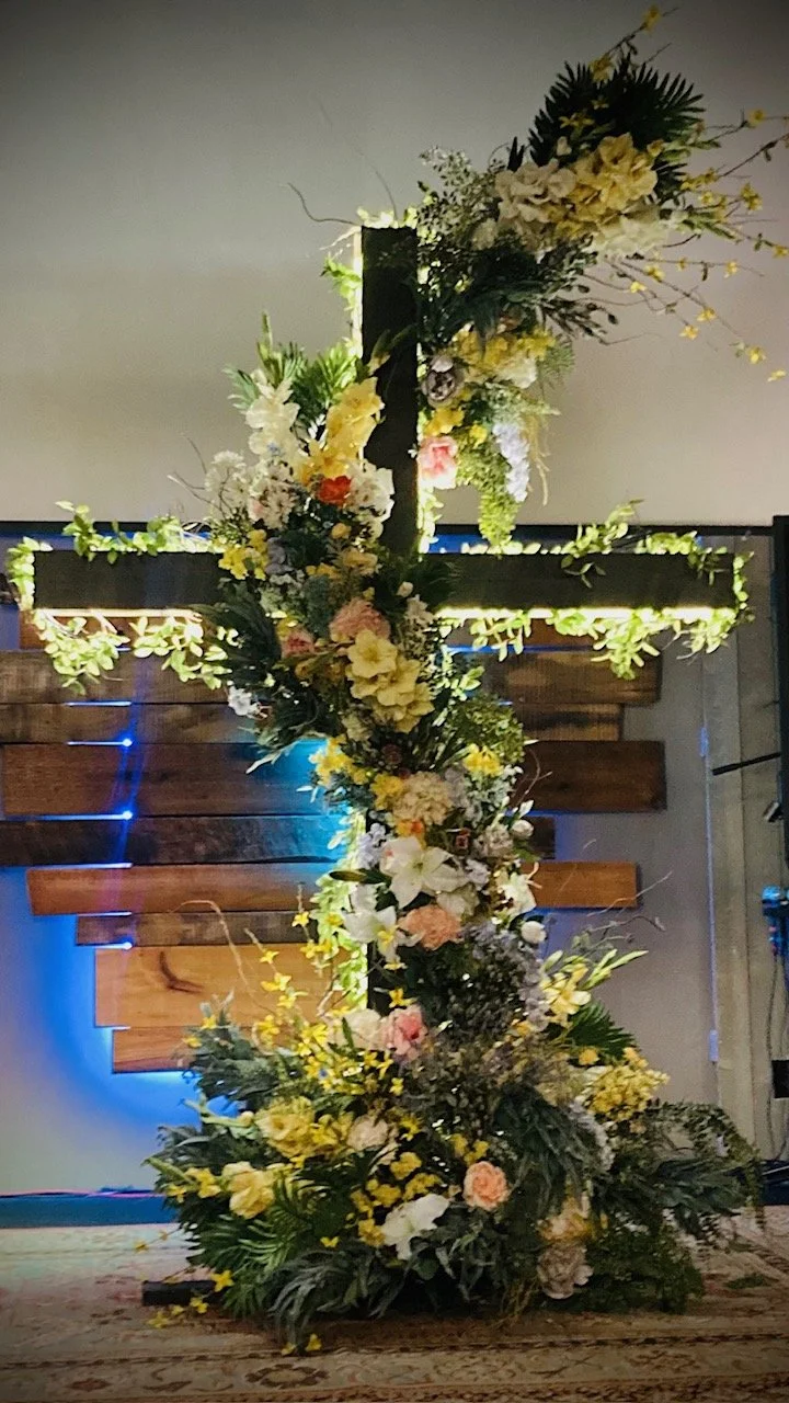 A decorative cross made of wood, adorned with flowers and greenery, standing indoors on a wooden floor.