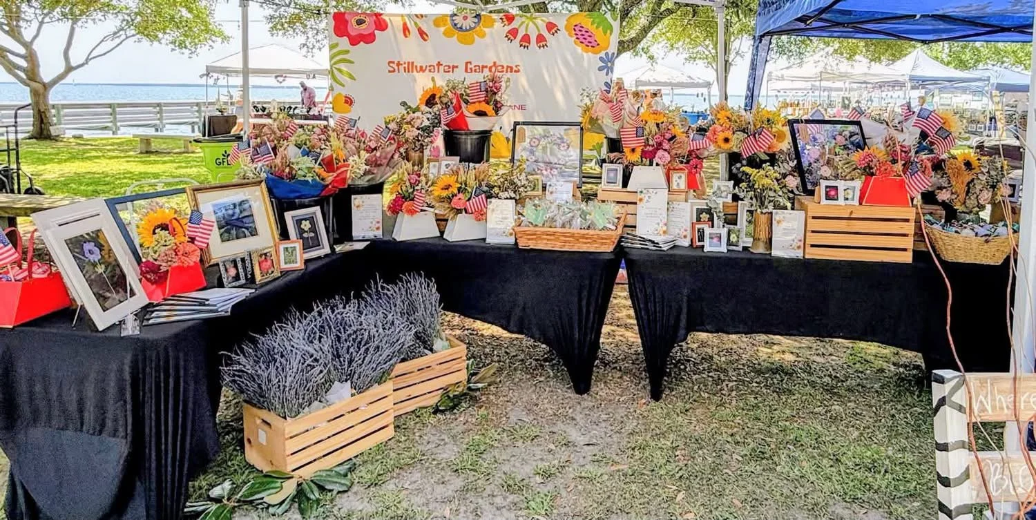 A display of flower arrangements, framed photographs, and American flags at Stillwater Gardens outdoor booth, with trees and a pool in the background.