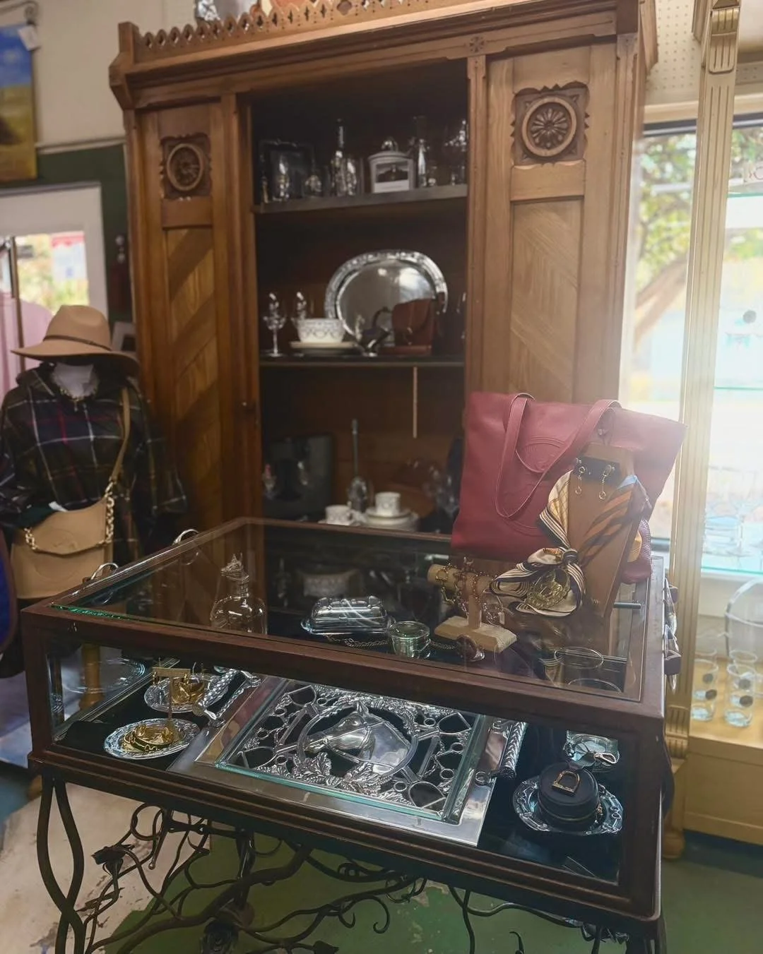A display of vintage and decorative items, including a large wooden cabinet with glass doors, a glass display case with jewelry and small collectibles, a mannequin dressed in a plaid shirt, scarf, and beige hat, and a red handbag with accessories, al