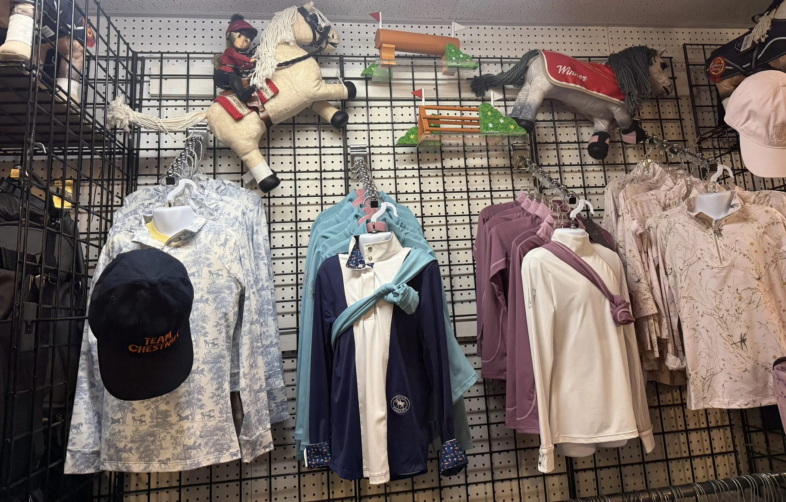Display of various clothing items and plush horses on a retail store wall. The clothing includes shirts, hoodies, and jackets on hangers, with some items labeled as winners or part of a team. Visitors can see hats, caps, and plush horse toys mounted 