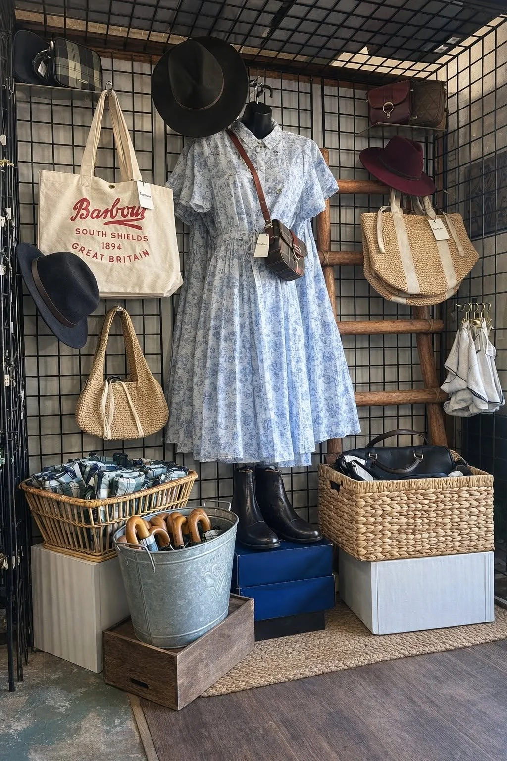 Vintage store display featuring a long, light blue floral dress on a mannequin, paired with black boots, surrounded by handbags, hats, a tote bag, umbrellas, and storage baskets on a black metal grid backdrop.