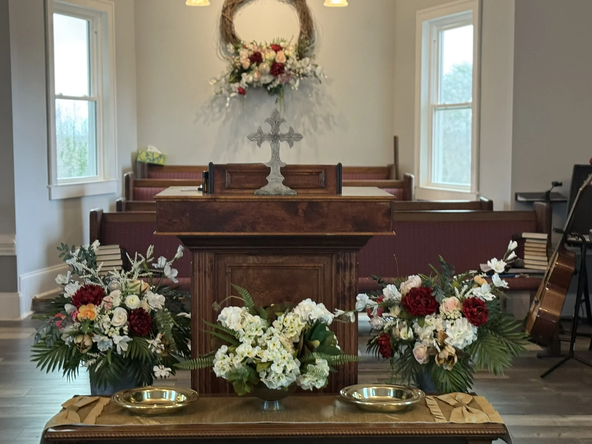 A church altar decorated with an arrangement of various colorful flowers, including roses and greenery, placed on a wooden table. Behind the altar, there is a cross and a wreath with more flowers hanging on the wall. The room has large windows allowi