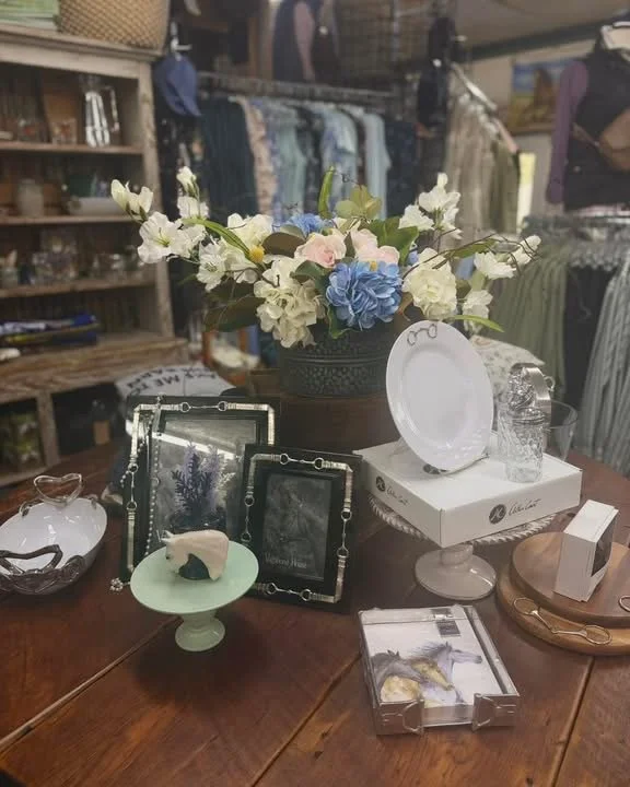 Decorative items arranged on a wooden table, including a floral arrangement in a vase, framed photos, jewelry displays, a white cake stand with a plate, and other small decorative objects.