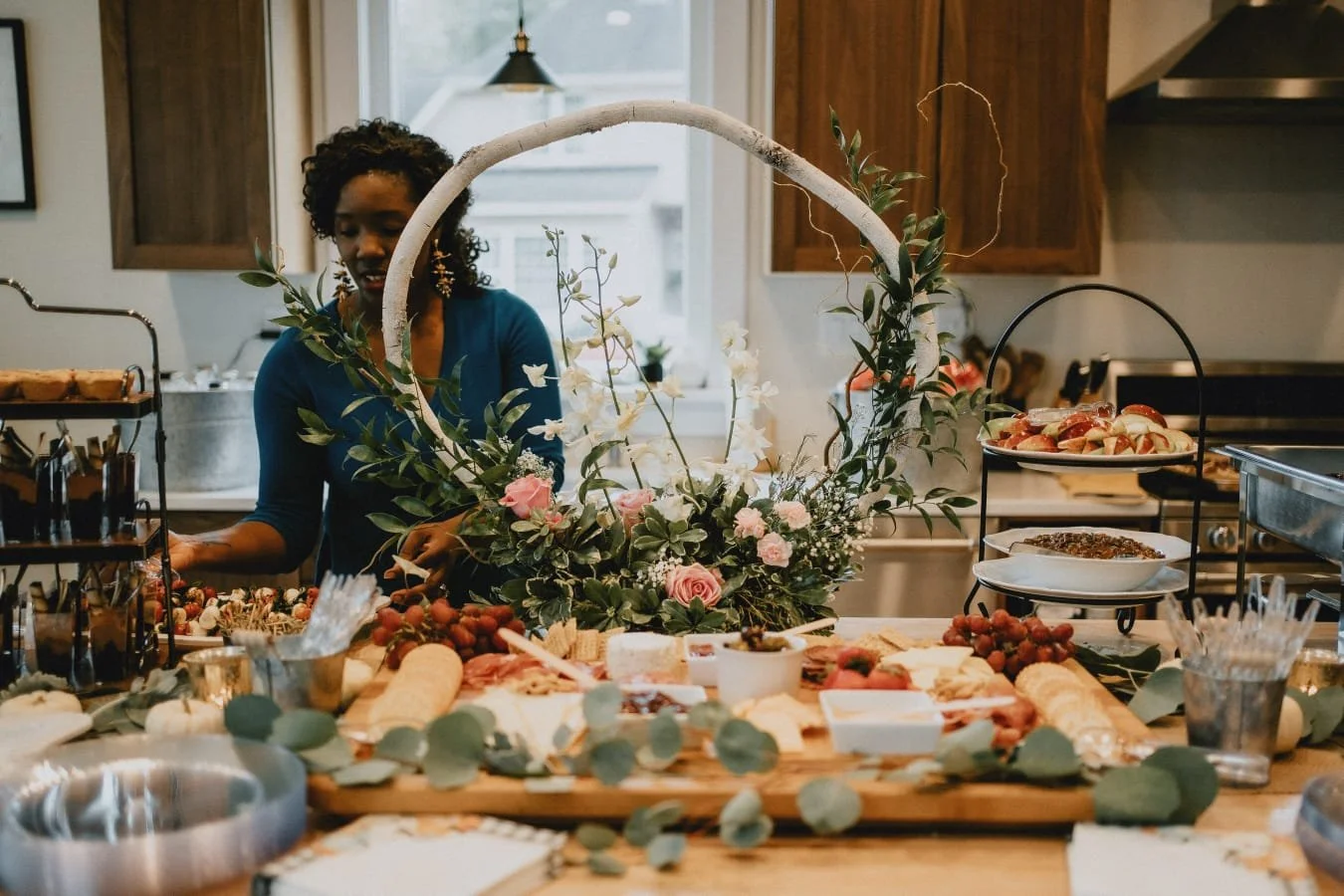 Woman arranging a charcuterie board decorated with flowers and greenery in a kitchen