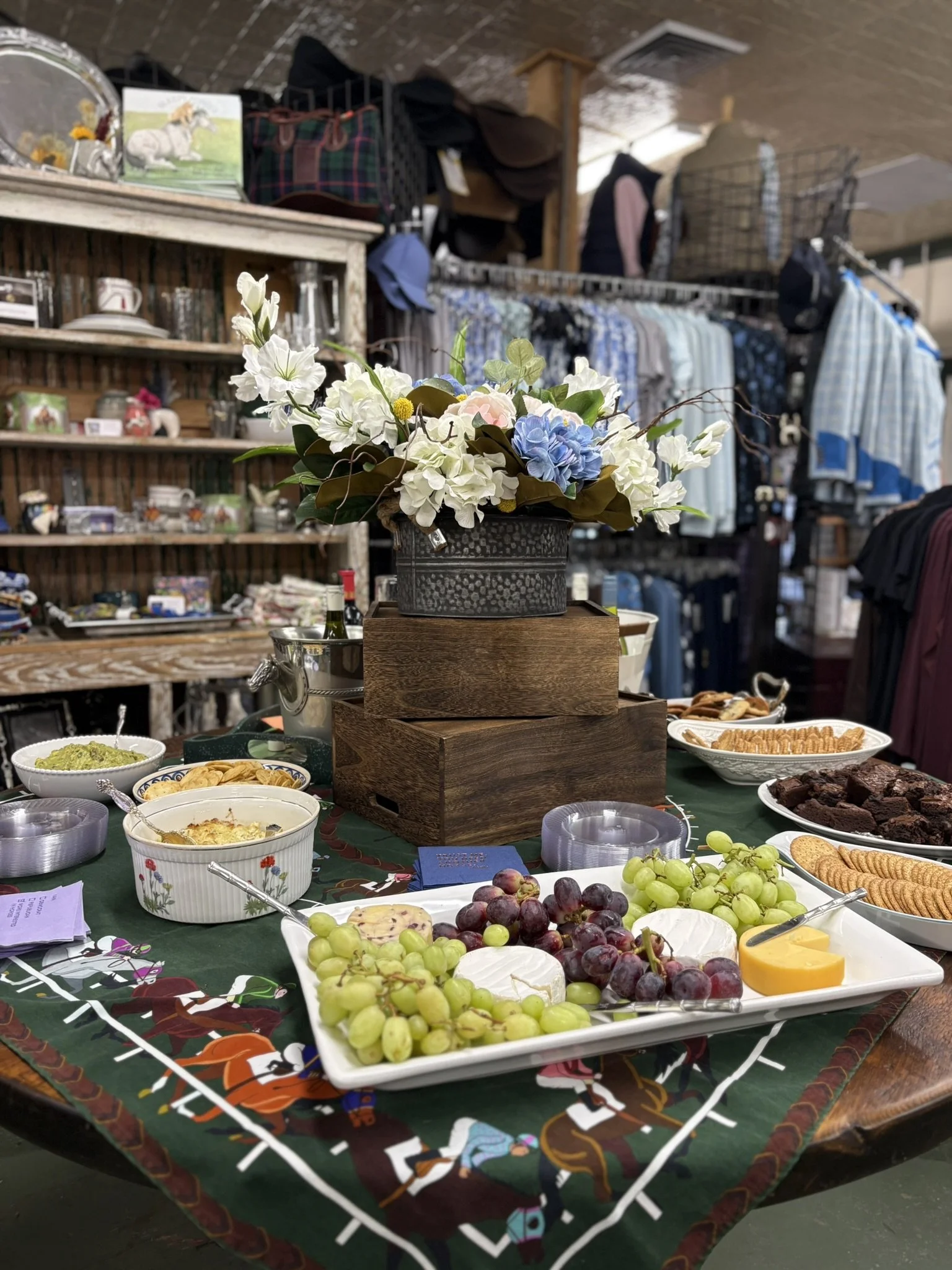 A table with various foods including grapes, cheese, crackers, brownies, and dips, decorated with a floral arrangement in a rustic container. In the background, there are shelves with collectibles, clothing racks, and people shopping in a store.