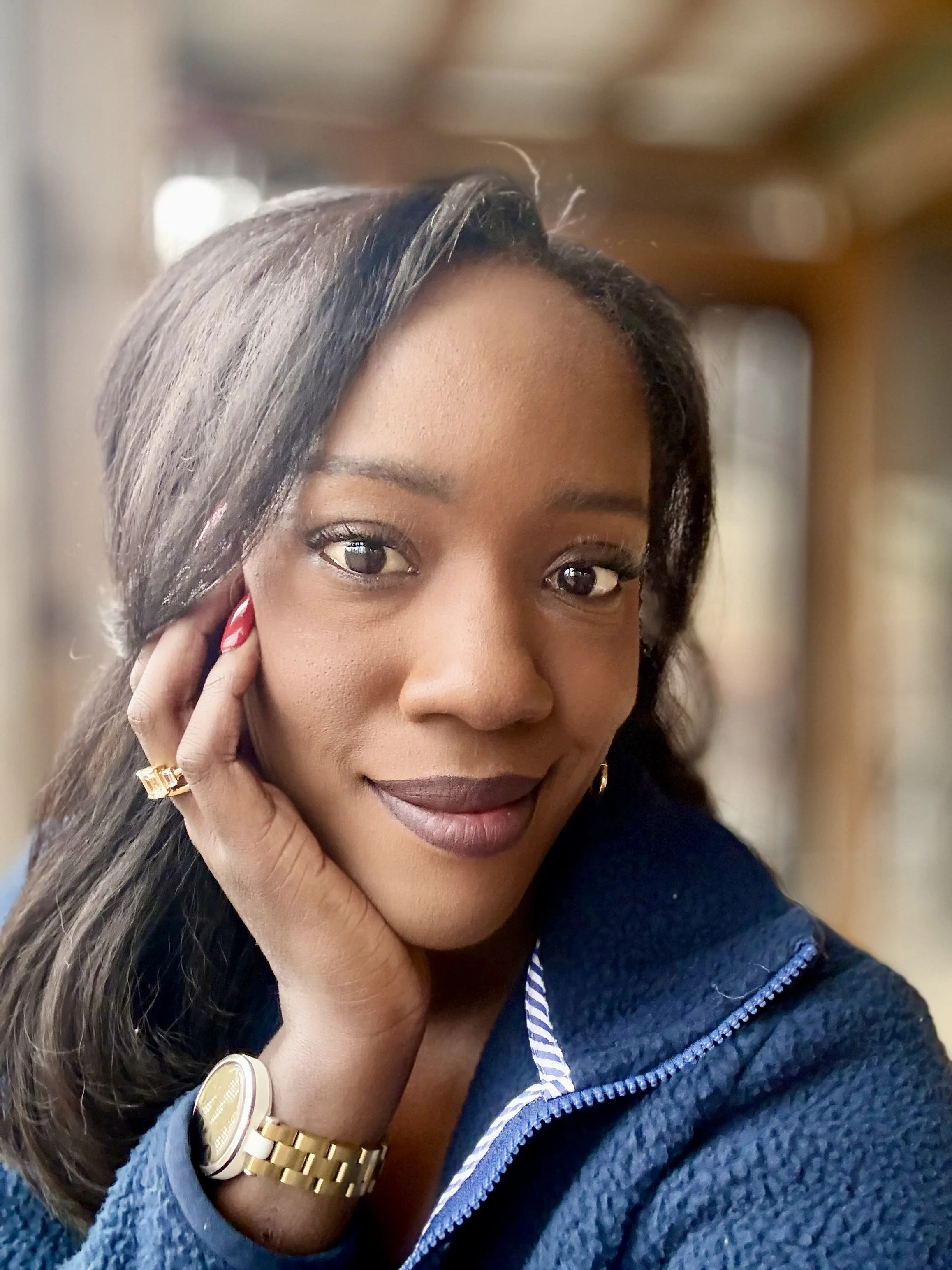 Close-up of a woman with medium-dark skin, dark straight hair, wearing a blue zip-up jacket, gold watch, and gold ring, resting her chin on her hand.