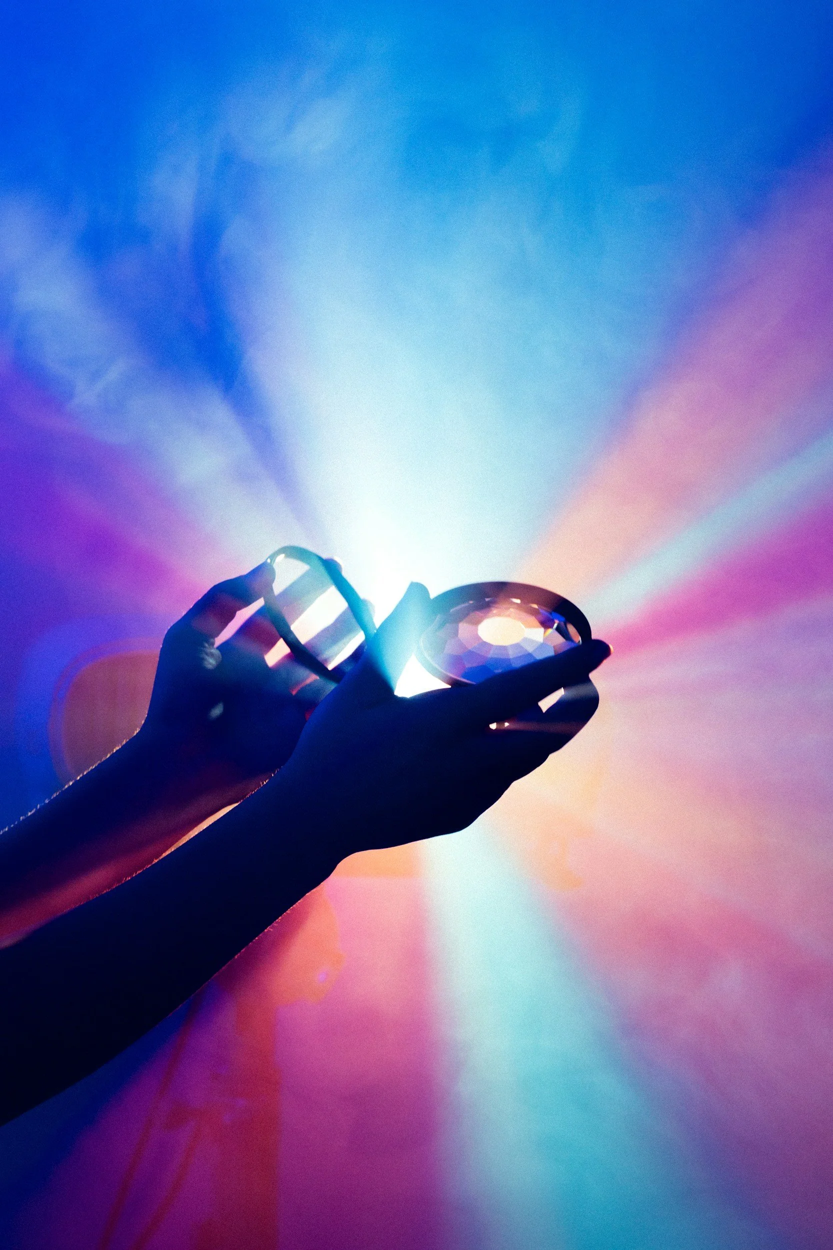 Silhouetted hand holding a faceted gemstone against a colorful blue, purple, and pink light background