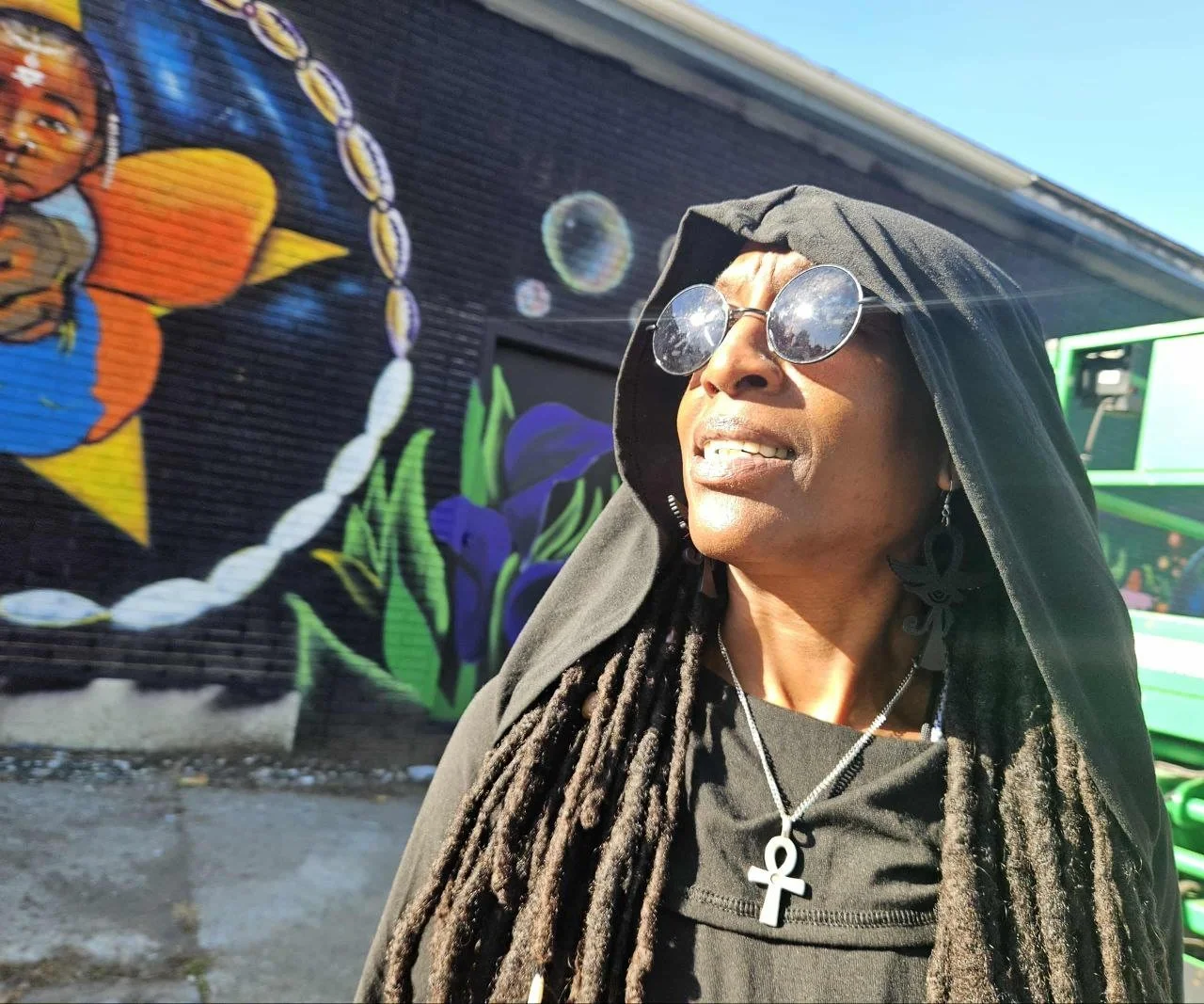 Black woman wearing a hood, round sunglasses, and an ankh necklace in front of a colorful mural