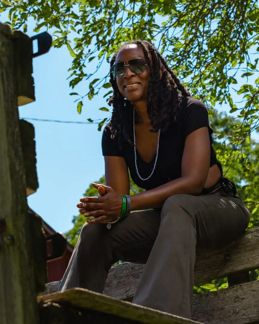 Smiling Black woman with long dreadlocks and sunglasses sitting outdoors on a wooden structure beneath leafy trees