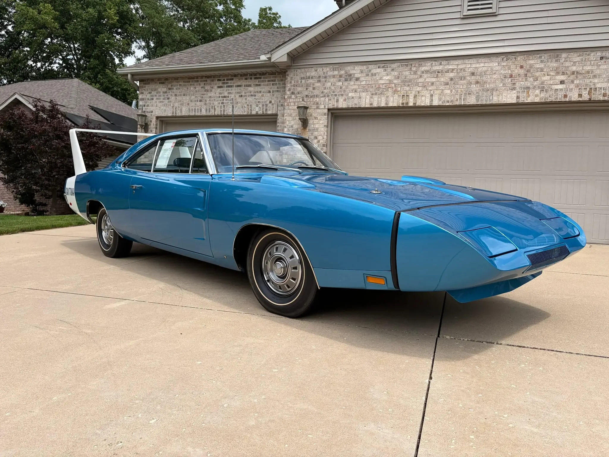 A blue classic sports car in a driveway of a suburban house, with a brick wall and garage doors in the background.