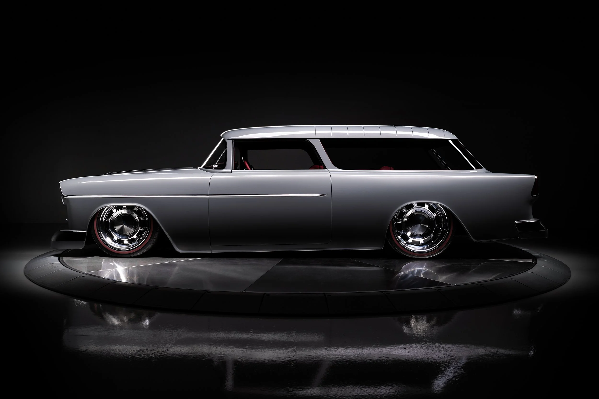 A sleek, silver vintage car with low profile and rounded edges, displayed in a professional studio setting on a reflective black floor with a dark background.