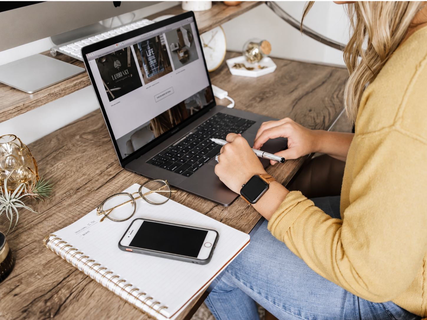 Woman browsing a website on a laptop at a wooden desk with a notebook, smartphone, and glasses nearby.