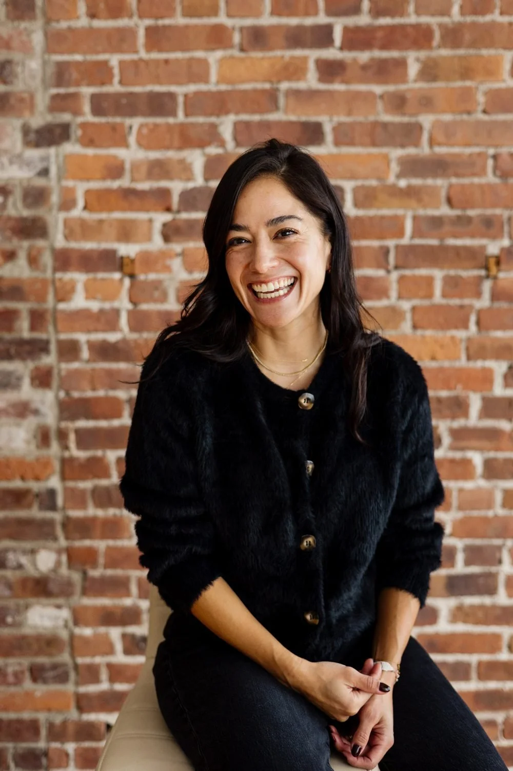 A smiling woman with dark hair wearing a black fuzzy sweater sitting in front of a brick wall.