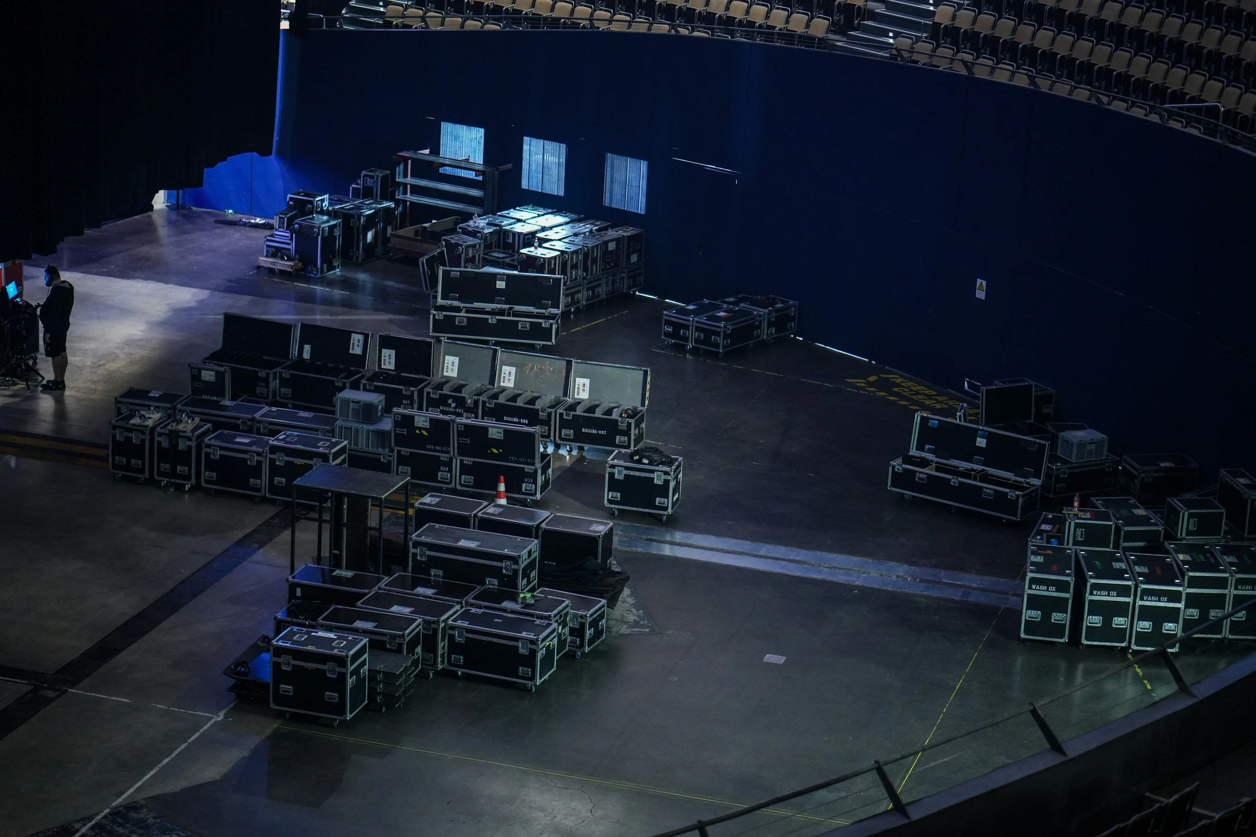 Stage setup with numerous black equipment cases and a person wearing a mask on the left side, in an auditorium or theater.