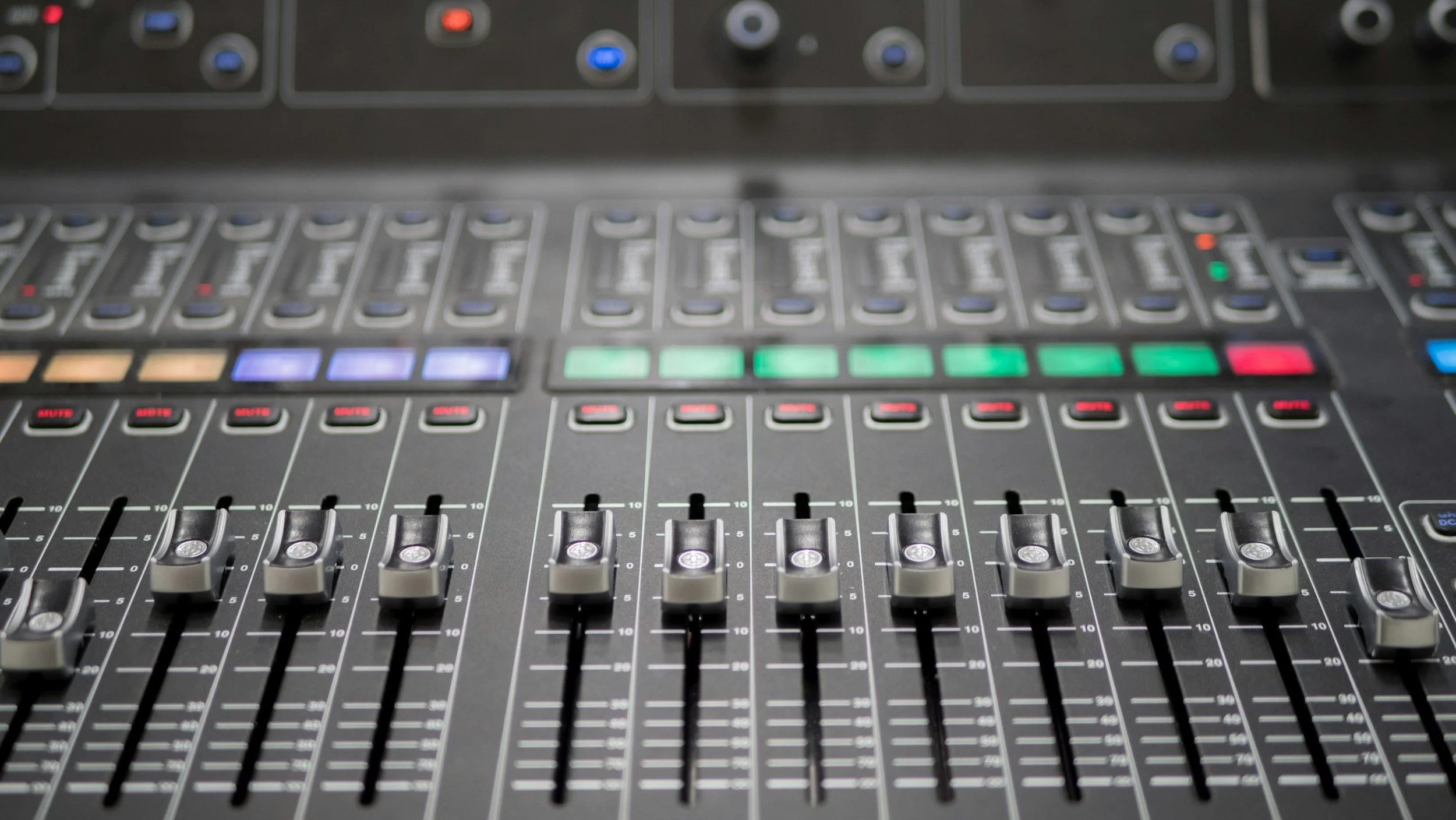 Close-up of an audio mixing console with faders, knobs, and colored buttons.