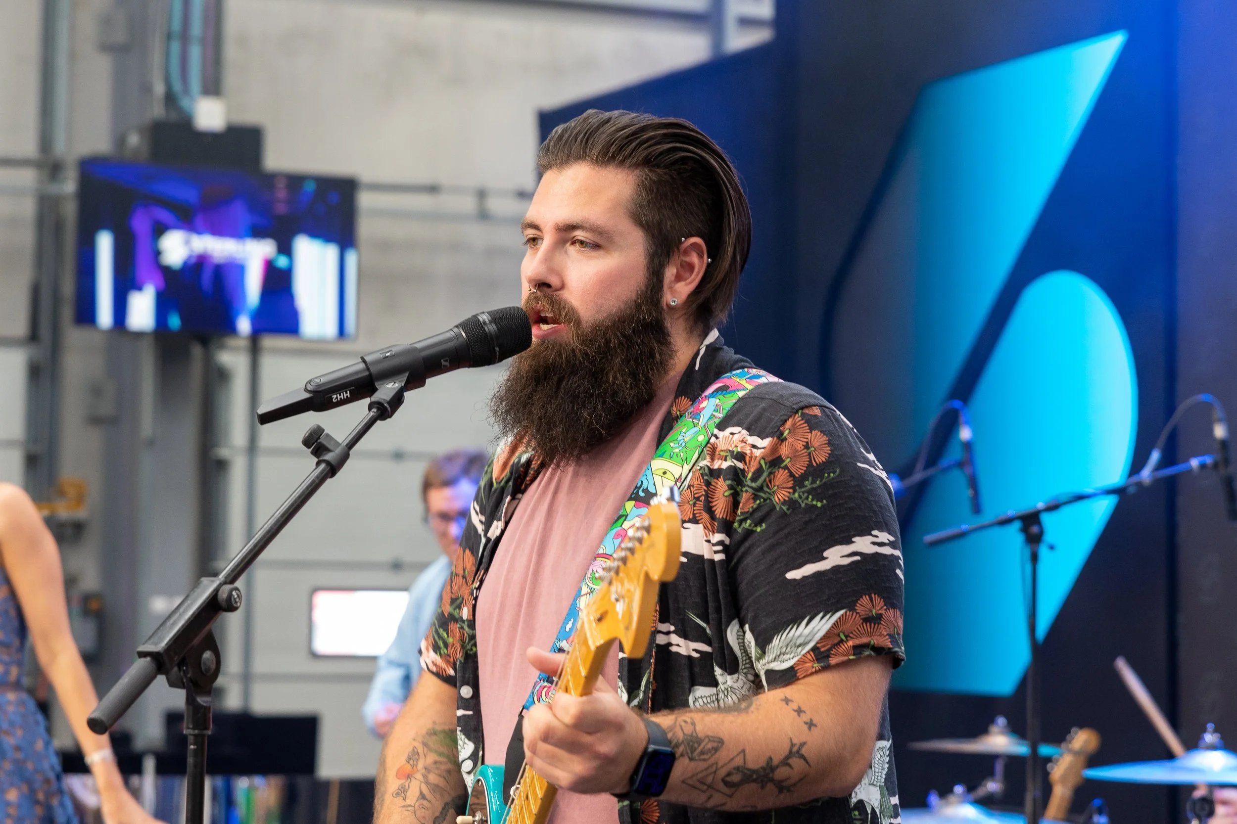 A man with a beard, tattoos, and styled hair is singing into a microphone while playing an electric guitar at an indoor event or performance.