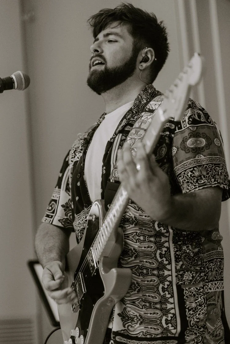 A young man with a beard and earrings playing an electric guitar during a performance, with a microphone nearby.