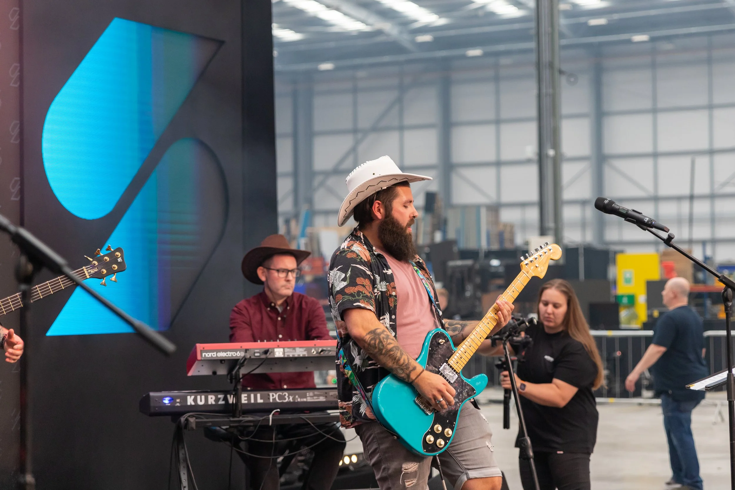 A man with a beard, wearing a cowboy hat, a floral shirt, and ripped jeans, playing an electric guitar at an indoor music event. Behind him are other musicians and equipment, and a large digital screen displaying a blue and black graphic.