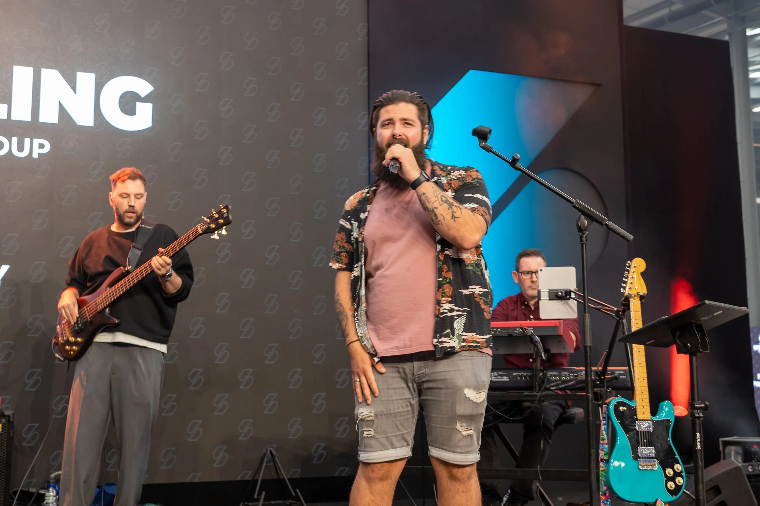 Musicians performing on stage, including a singer with a beard, long hair, and tattoos, holding a microphone, with a guitarist and keyboardist in the background, and guitars on stands.