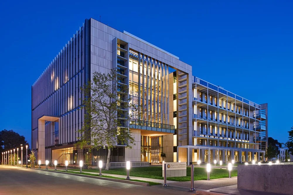 Modern university building with glass facade and illuminated windows at dusk, surrounded by pathway lights and green landscaping.